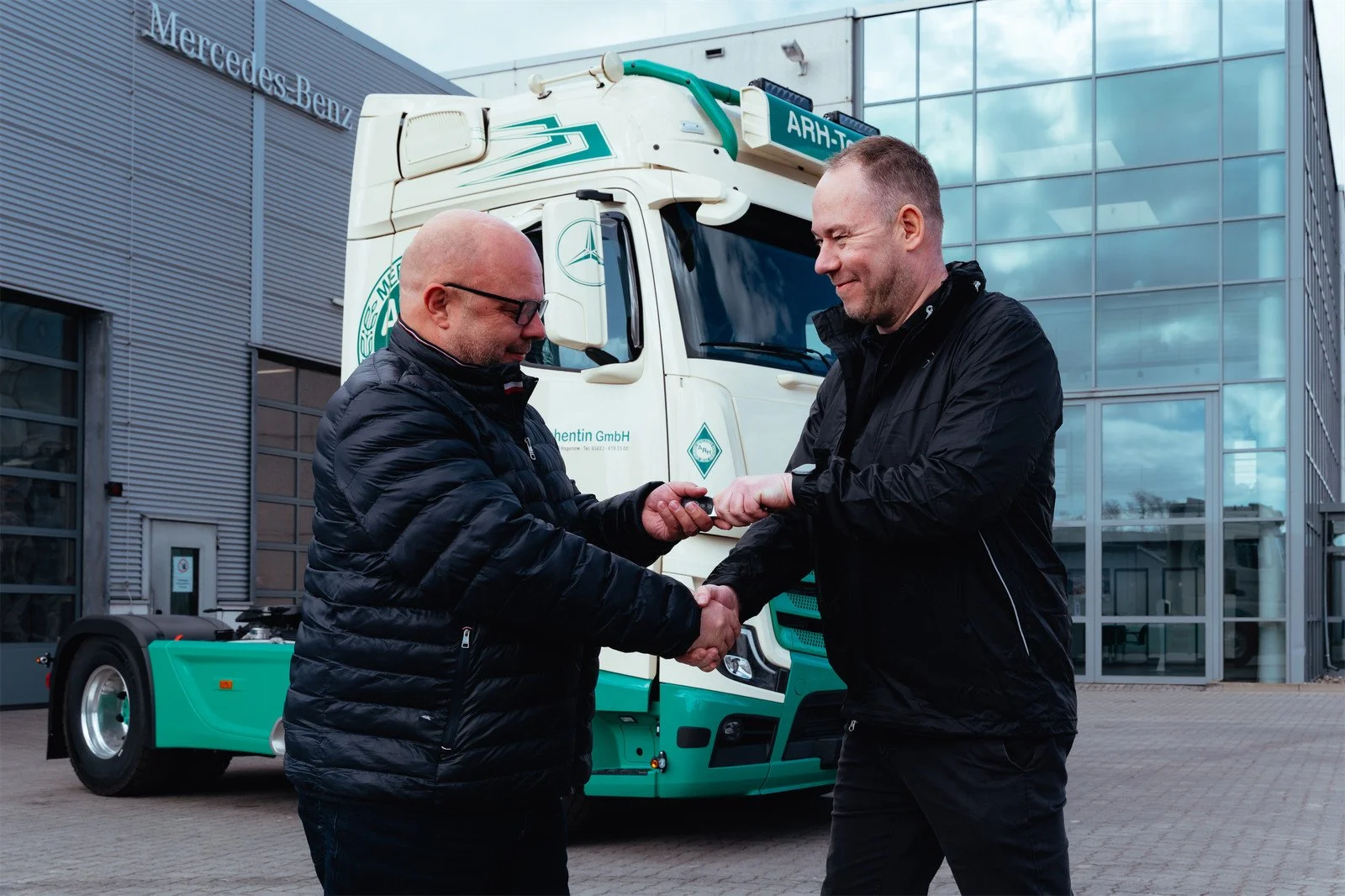 Zwei Männer tauschen einen Autoschlüssel vor einem Mercedes-Benz Transporter. Einer der Männer übergibt den Schlüssel dem anderen. Das Bild ist draußen vor einem modernen Gebäude mit Glasfassade aufgenommen.