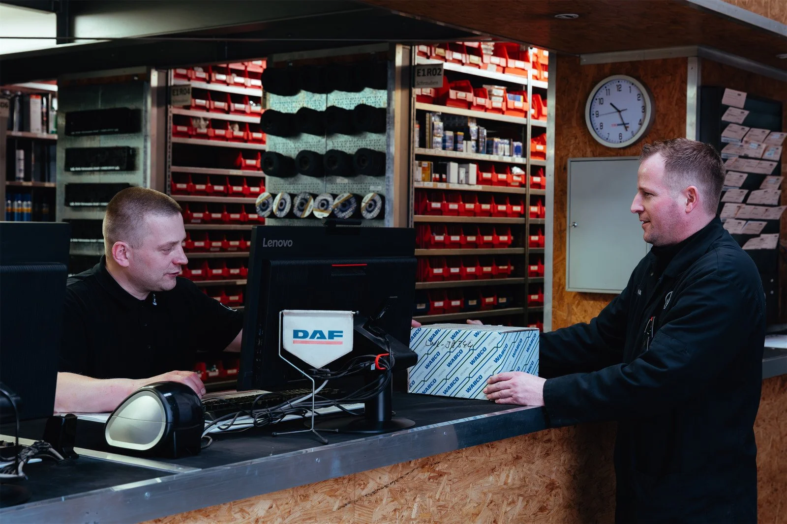 Zwei Männer in einer Werkstatt, einer übergibt dem anderen ein Paket mit einer WABCO- und WABCO-Logo. Im Hintergrund sind Werkstattregale mit roten Boxen und Werkzeugen.