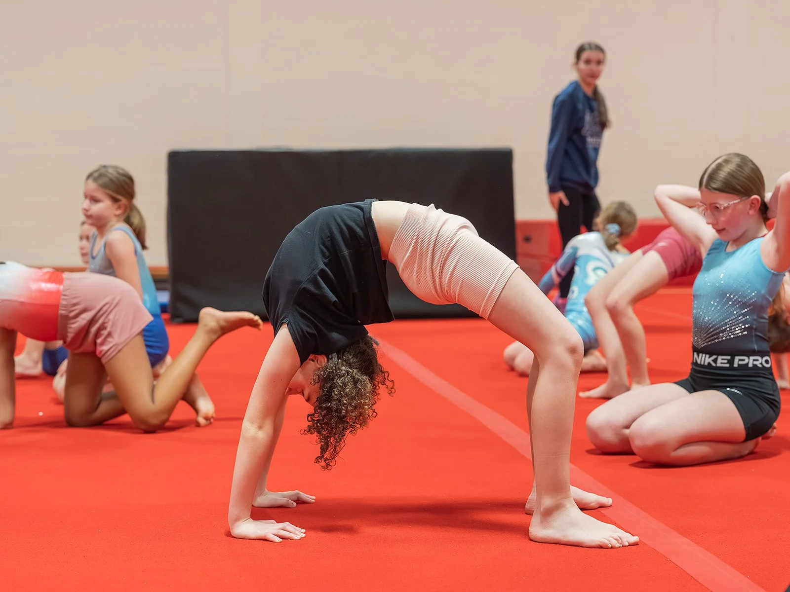 Children participating in a gymnastics class, stretching on a blue mat in a gym.