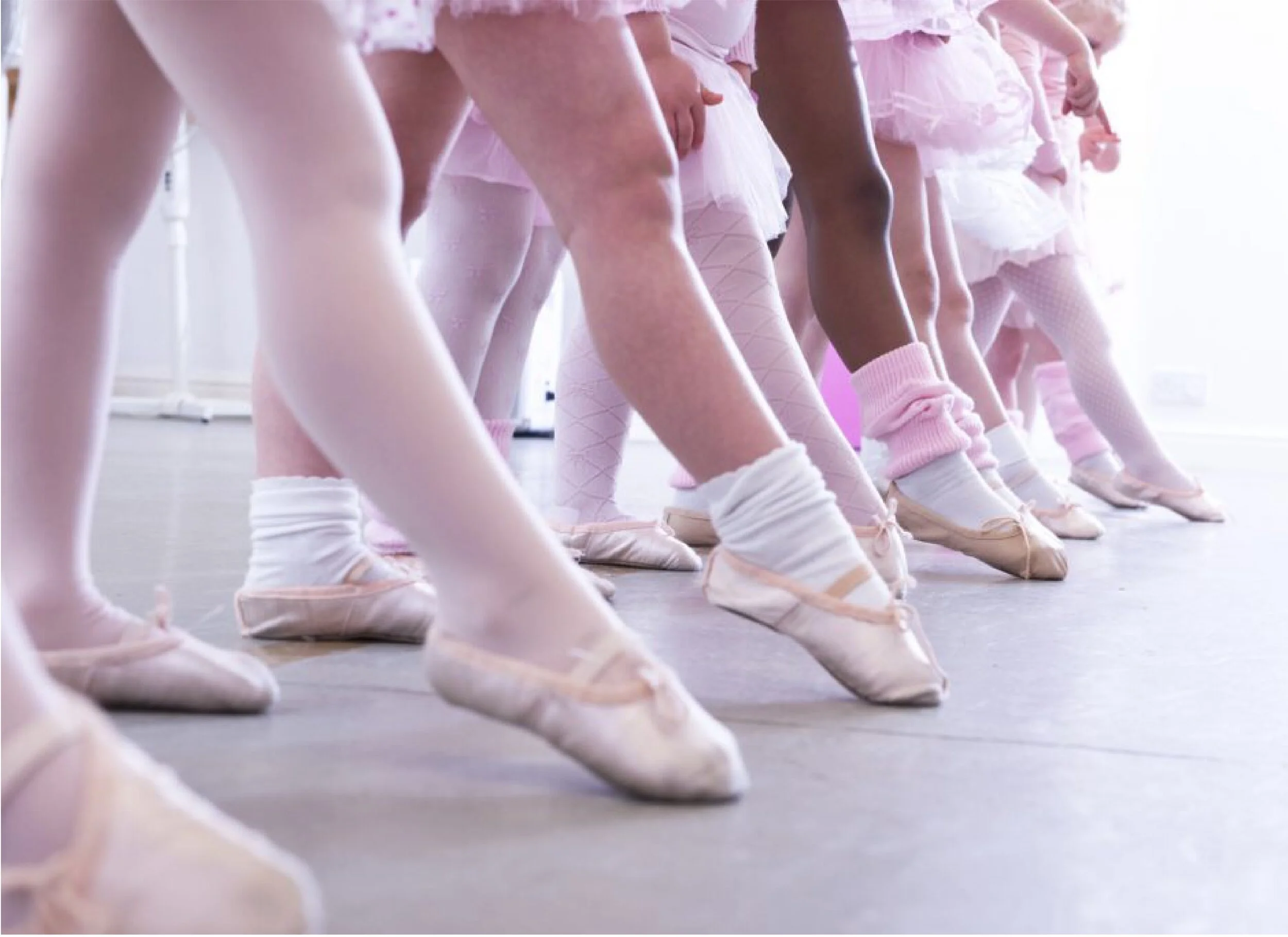 Children wearing ballet shoes and pastel clothing practicing dance positions on a studio floor.