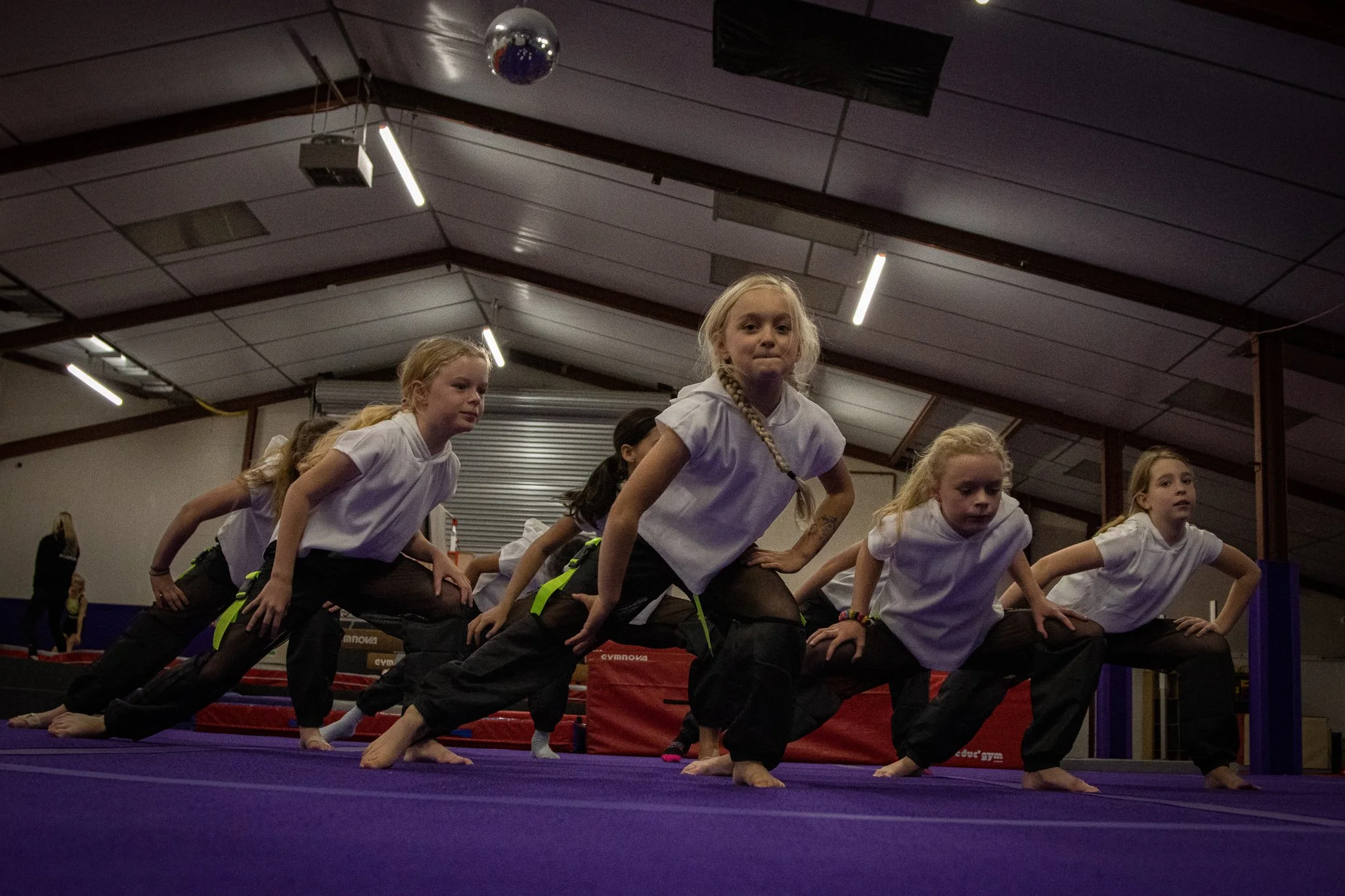 Children in a gymnasium practicing a dance routine on a purple mat, wearing white shirts and black pants, with a disco ball hanging from the ceiling.