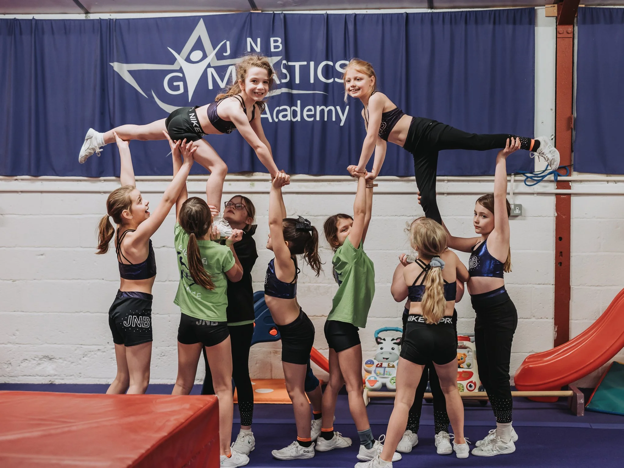 Group of children practicing acrobatics at a gymnastics academy, with two girls supported in the air by a group of others.