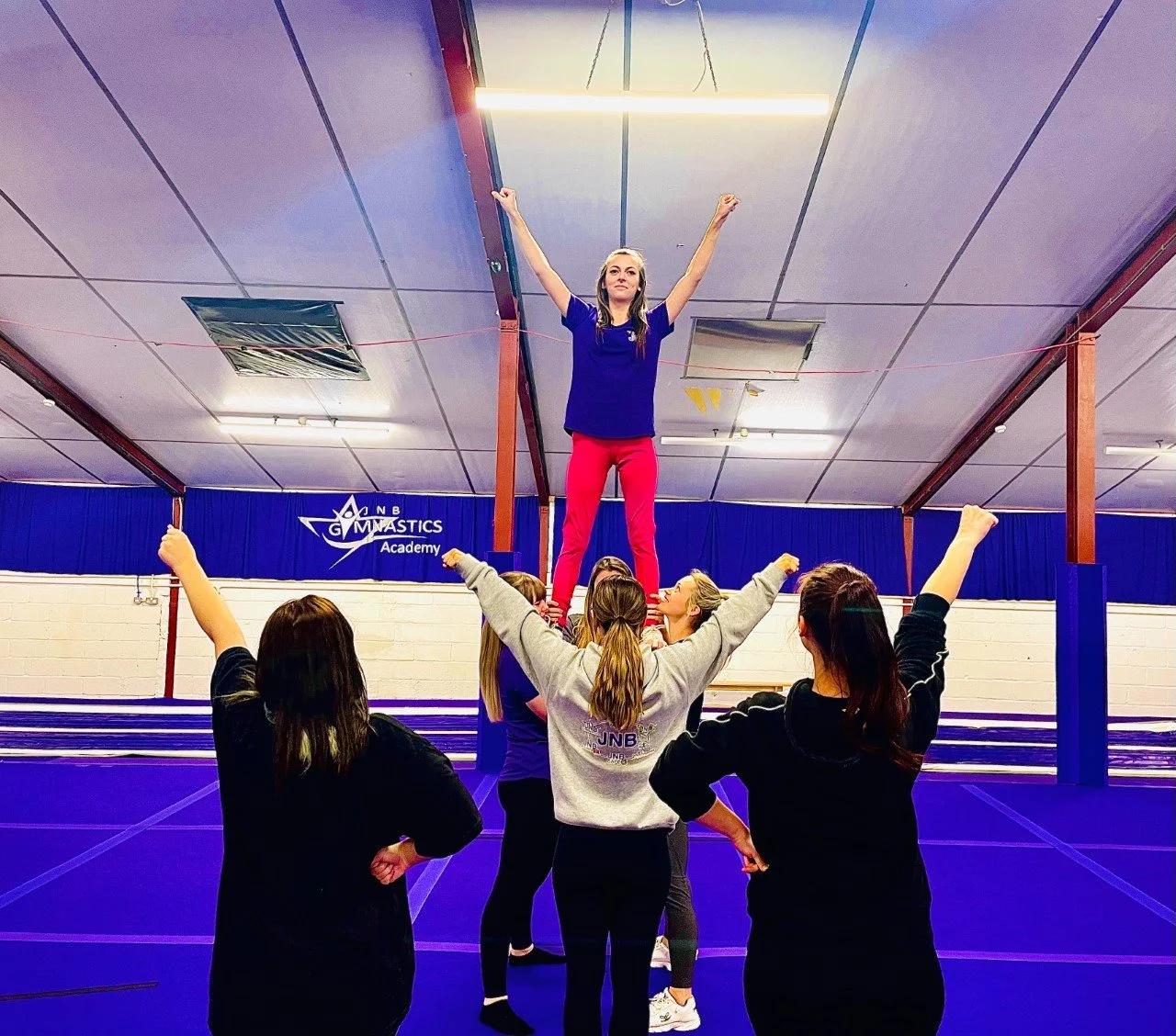 Cheerleaders practicing a stunt indoors at a gymnastics academy on a blue mat; one person is elevated with arms raised, supported by others.