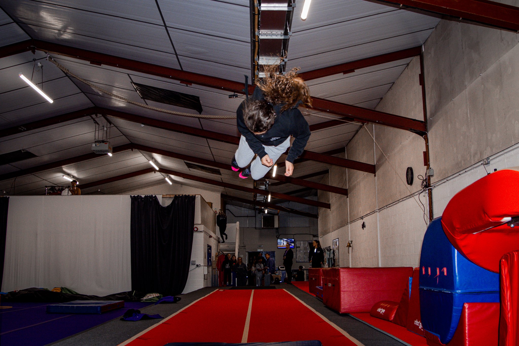 Person performing a flip on an indoor trampoline in a gymnasium with padded equipment and spectator background.