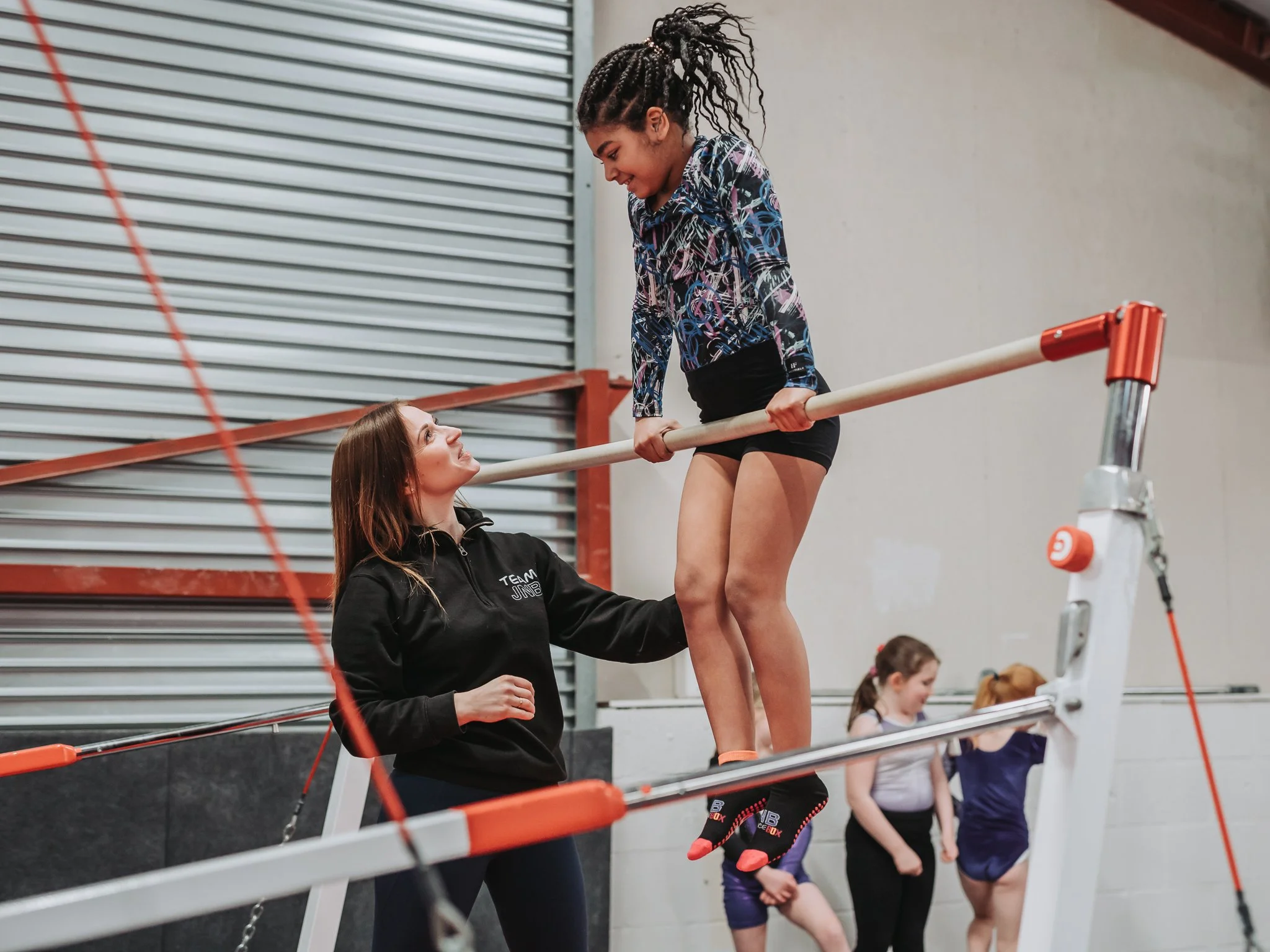 A young gymnast practicing on the uneven parallel bars with a coach's assistance in a gym. Other gymnasts are visible in the background.