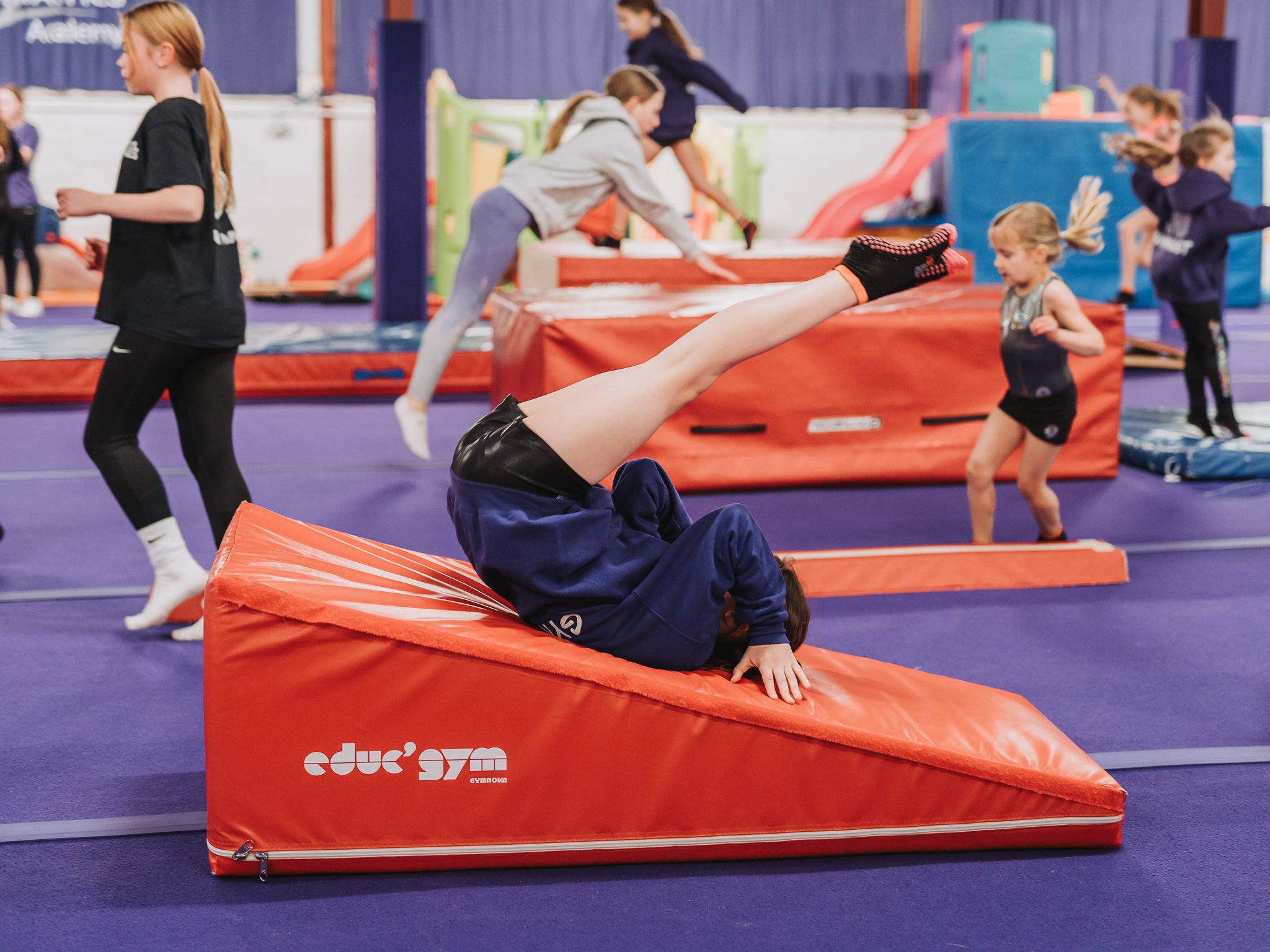 Children practicing gymnastics in a gymnasium with red padded equipment and a purple floor.