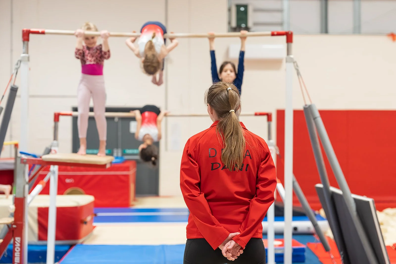Two young girls practicing gymnastics on an orange mat, one doing a split with arms up, the other leaning forward, both wearing blue leotards in a gymnasium.