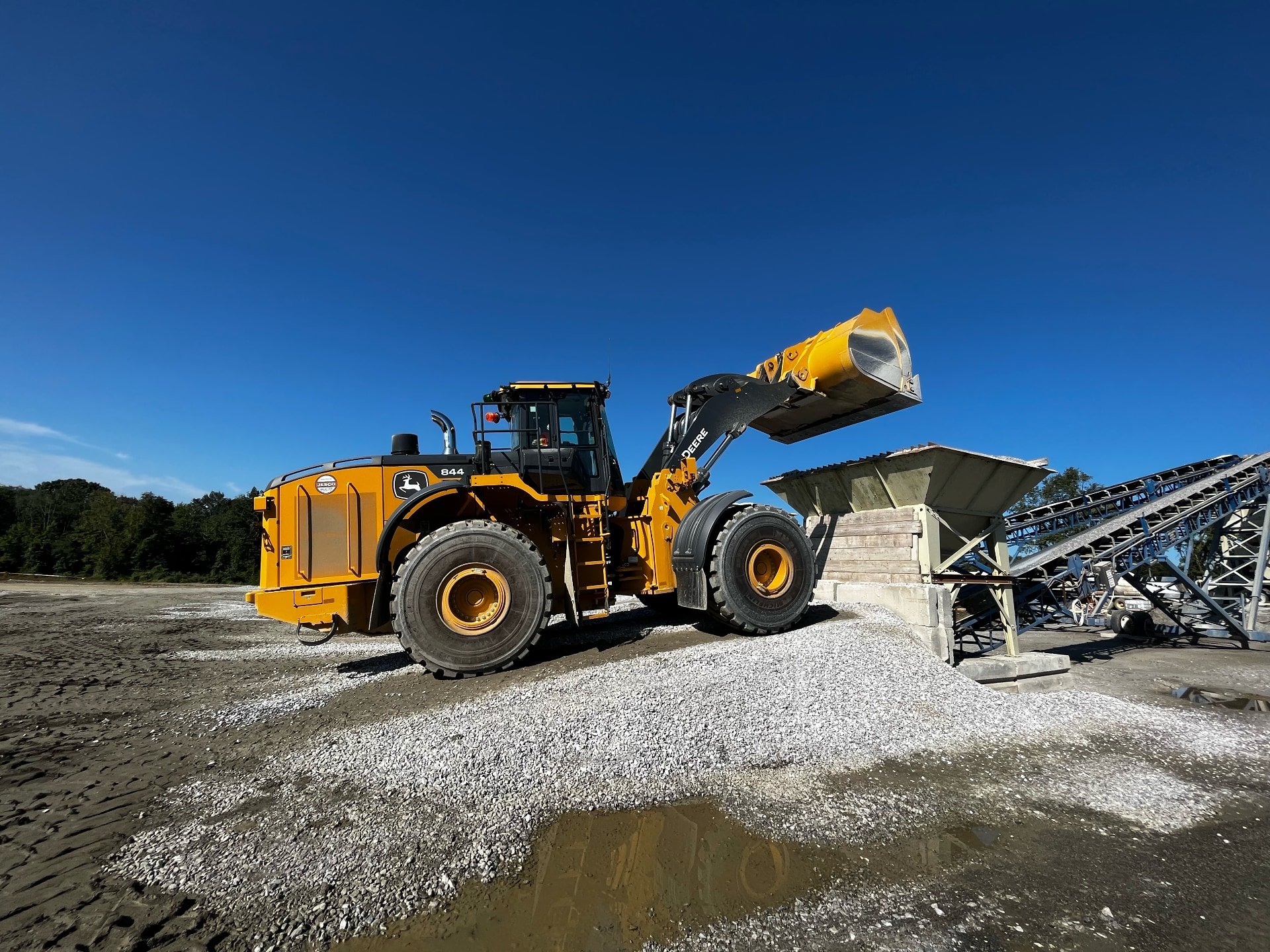 A large yellow bulldozer with a black cabin and the John Deere logo is moving gravel at a construction site under a clear blue sky.