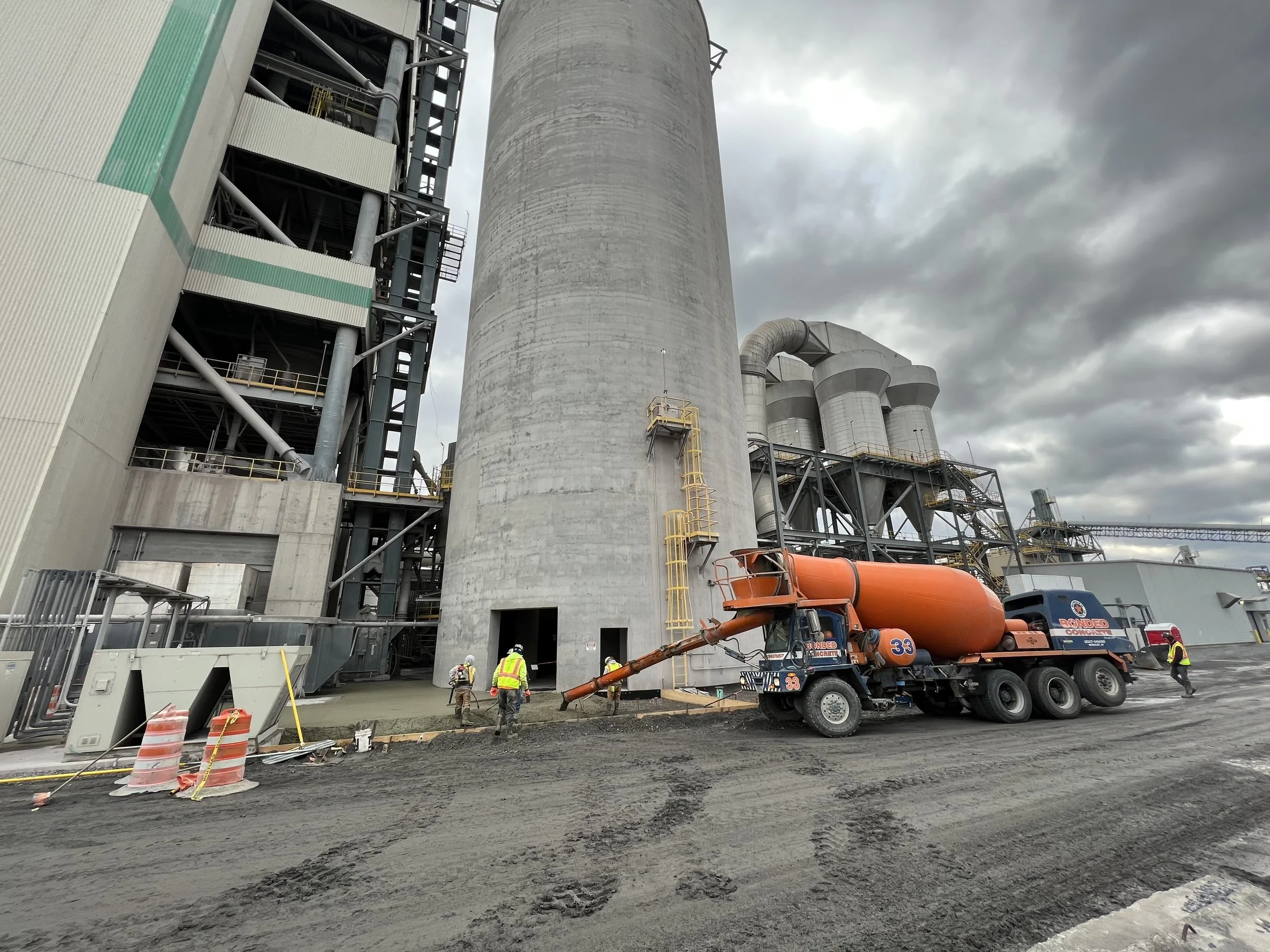 Construction site with workers and a cement mixer truck pouring concrete onto a surface next to large industrial structures under cloudy sky.