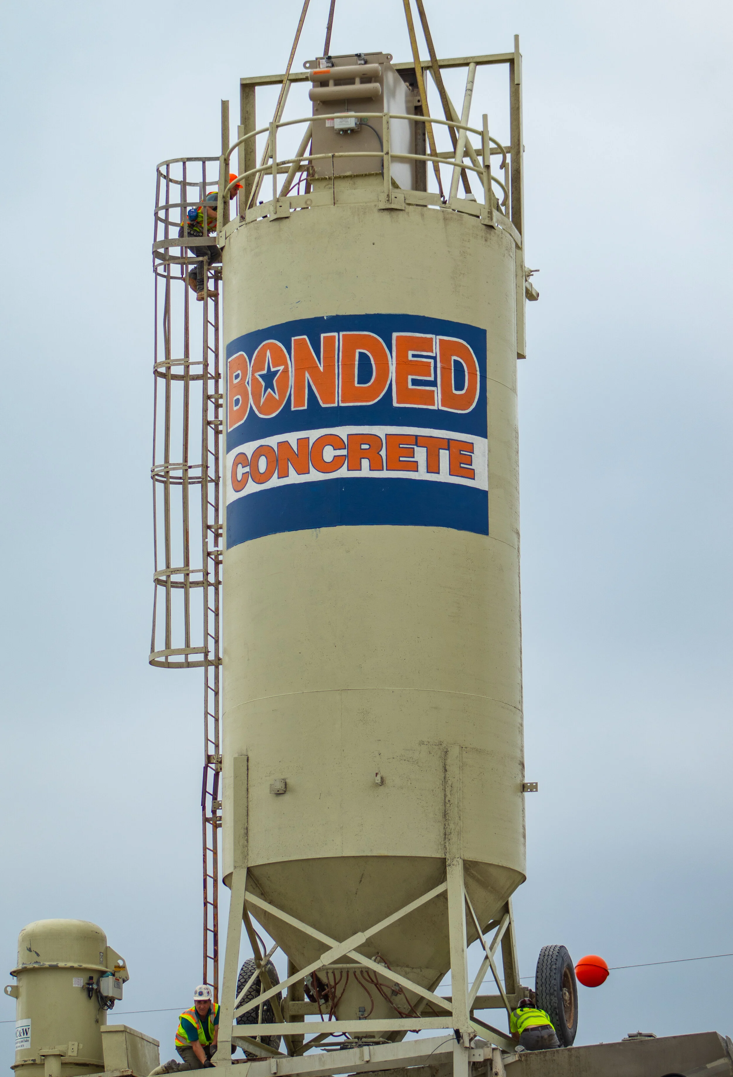 A large concrete silo labeled 'BONDED CONCRETE' with two workers in reflective vests and helmets working at the base of the silo.