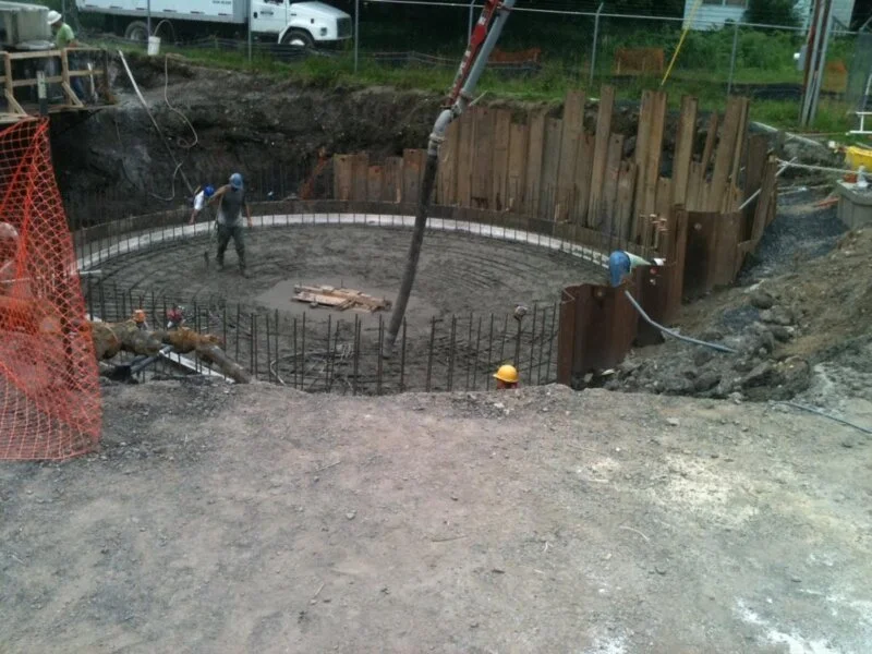 Construction site with a circular concrete foundation, steel reinforcement bars, and workers in safety gear, surrounded by dirt and construction materials.