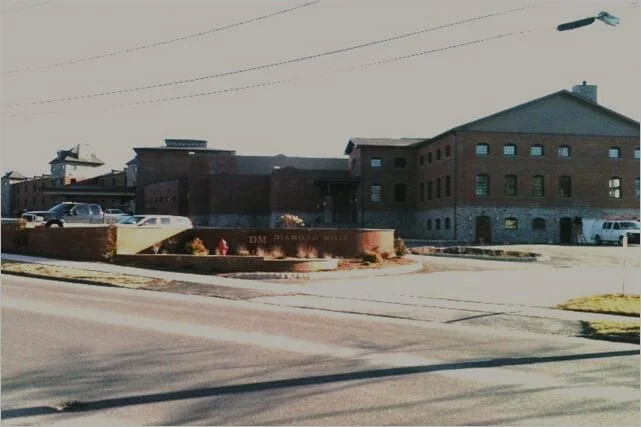 A brick building with multiple windows and a sign reading "The Old Courthouse Museum" in front. A street with cars parked along the side and a small landscaped area with flowers. Power lines and a streetlight are visible in the sky.