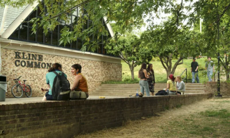 People gathered outside the Kline Common building in a park-like setting with trees and stairs, some sitting on the ledge, others standing and conversing.