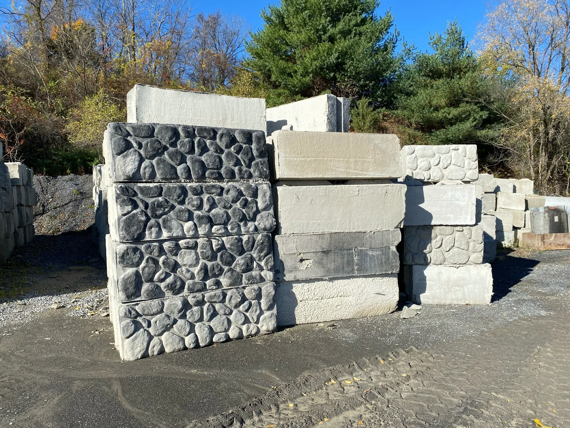 Stacked concrete blocks and stones at a construction supply yard with trees and blue sky in the background.