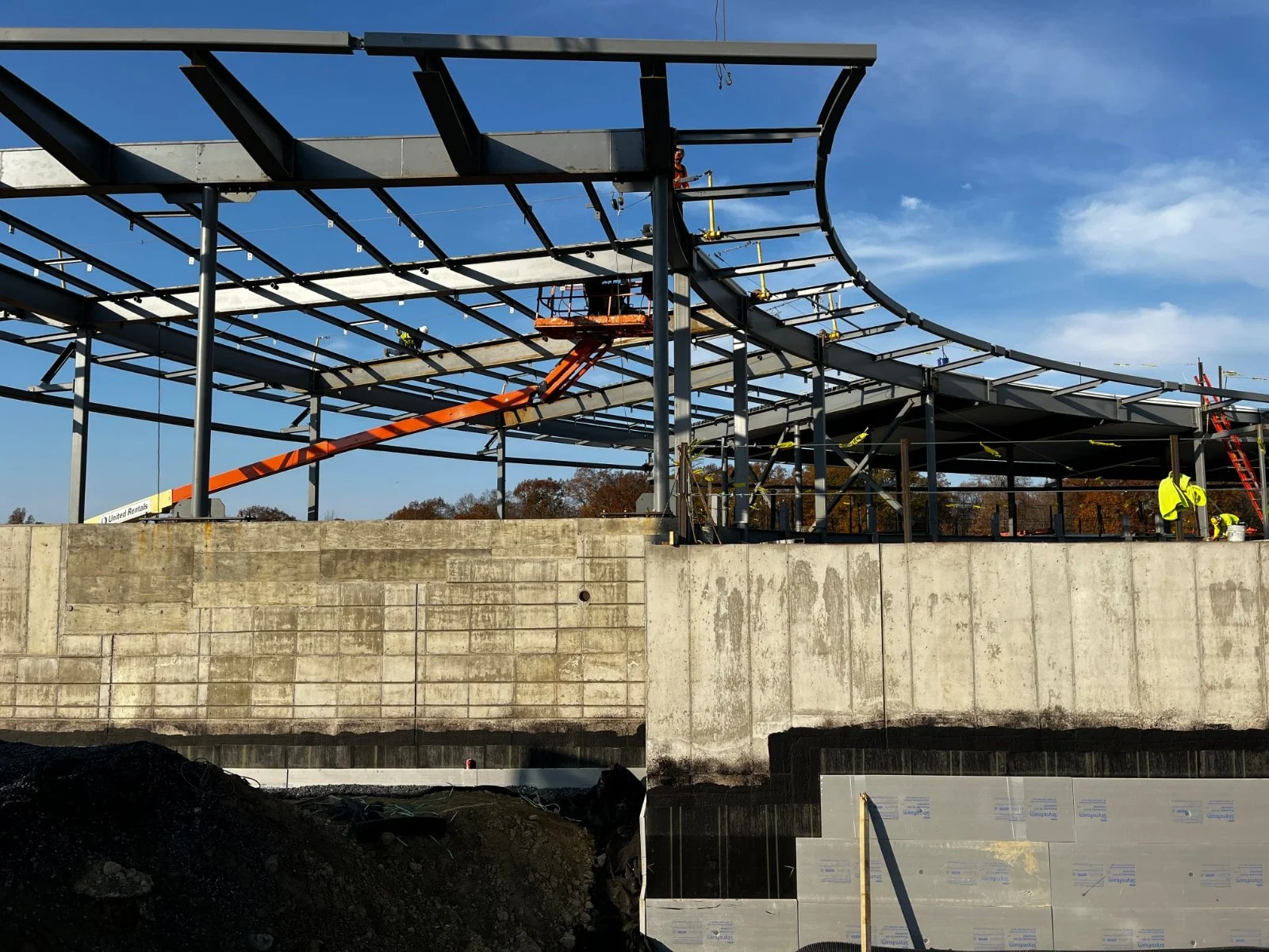 Construction workers building a large structure with metal framework on a concrete foundation under a blue sky.