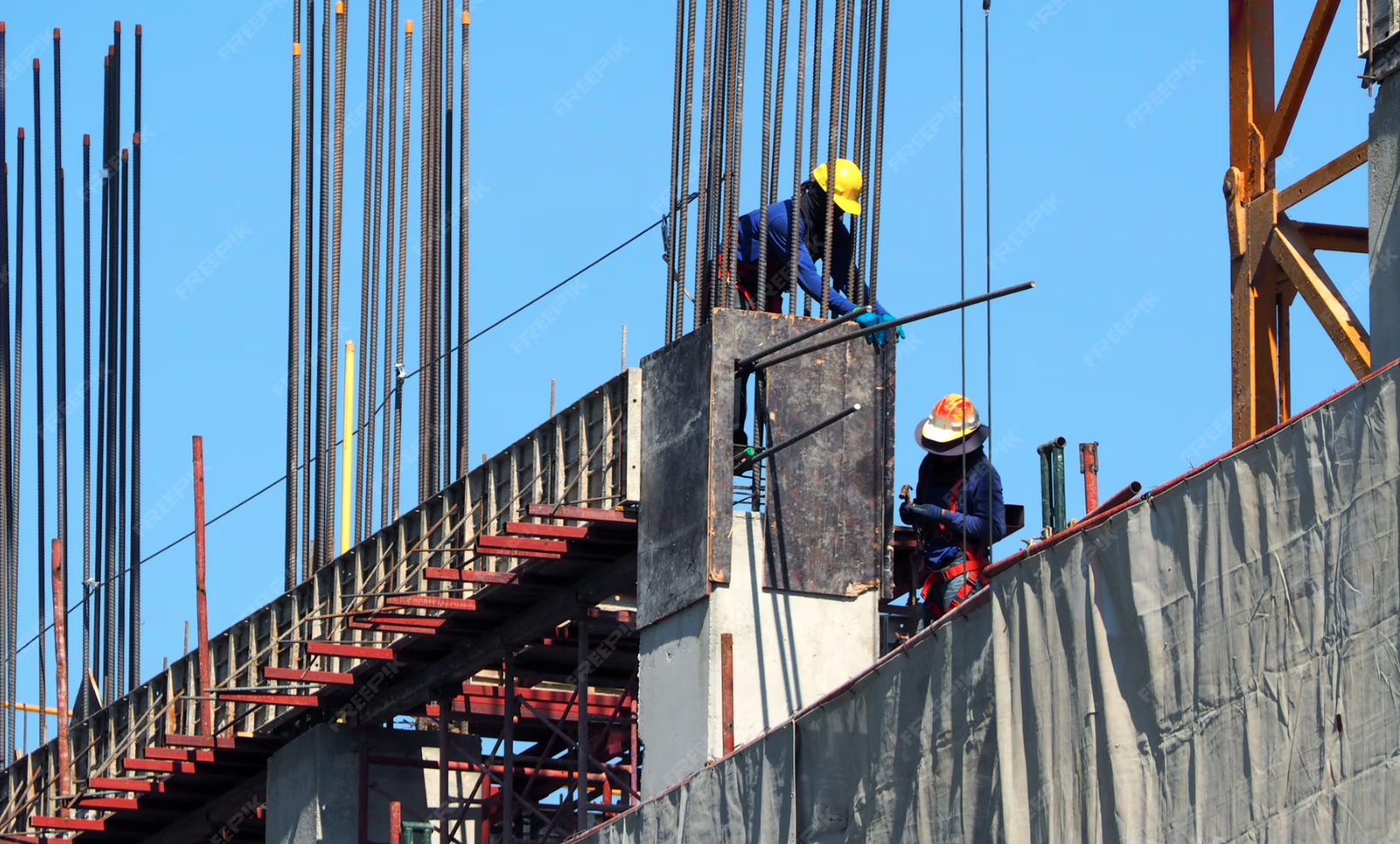 Construction workers in safety gear working on a building under construction with steel rebar and wooden scaffolding against a blue sky.