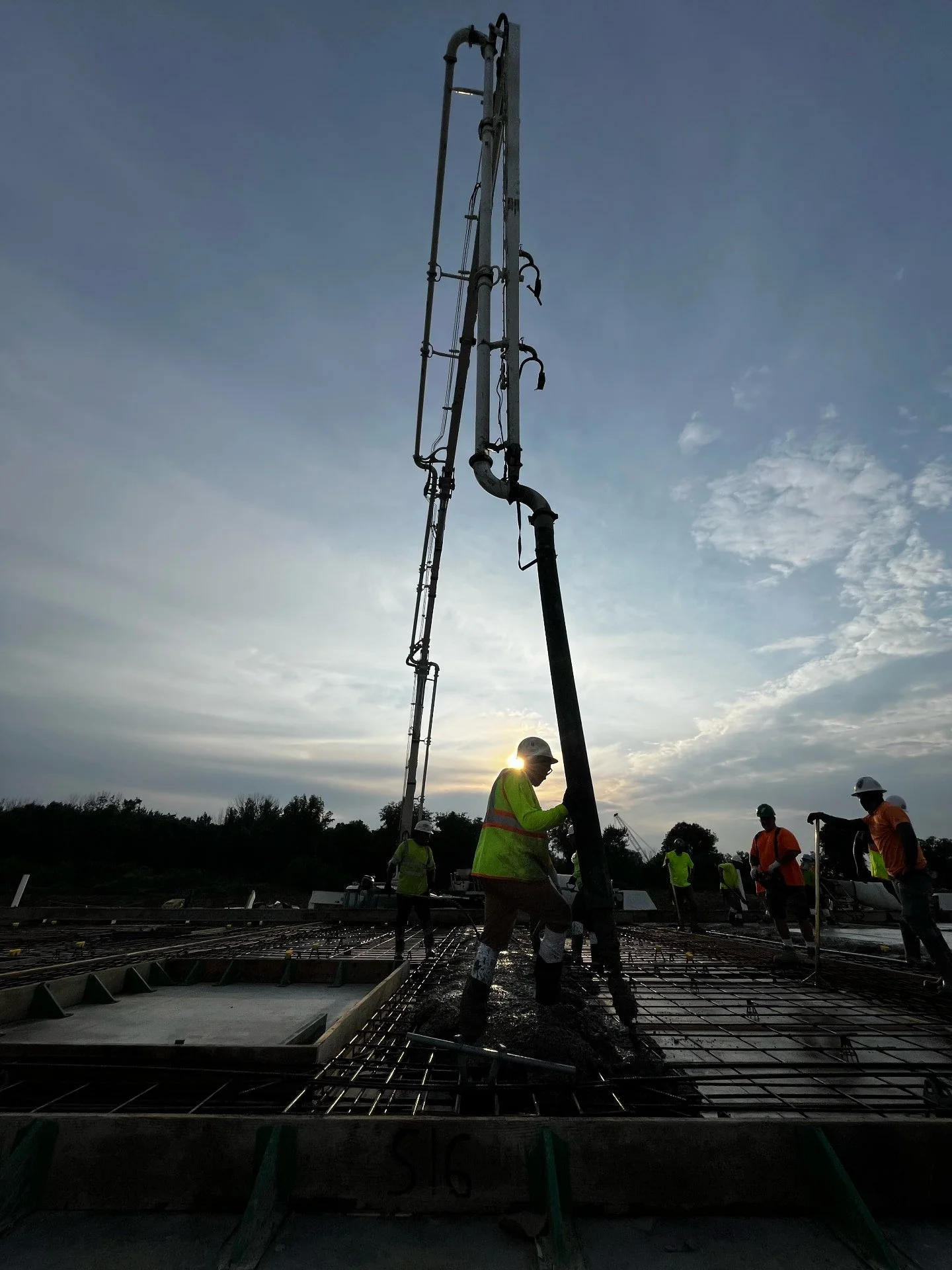 Construction workers building a concrete foundation with rebar, during sunset, wearing safety gear.