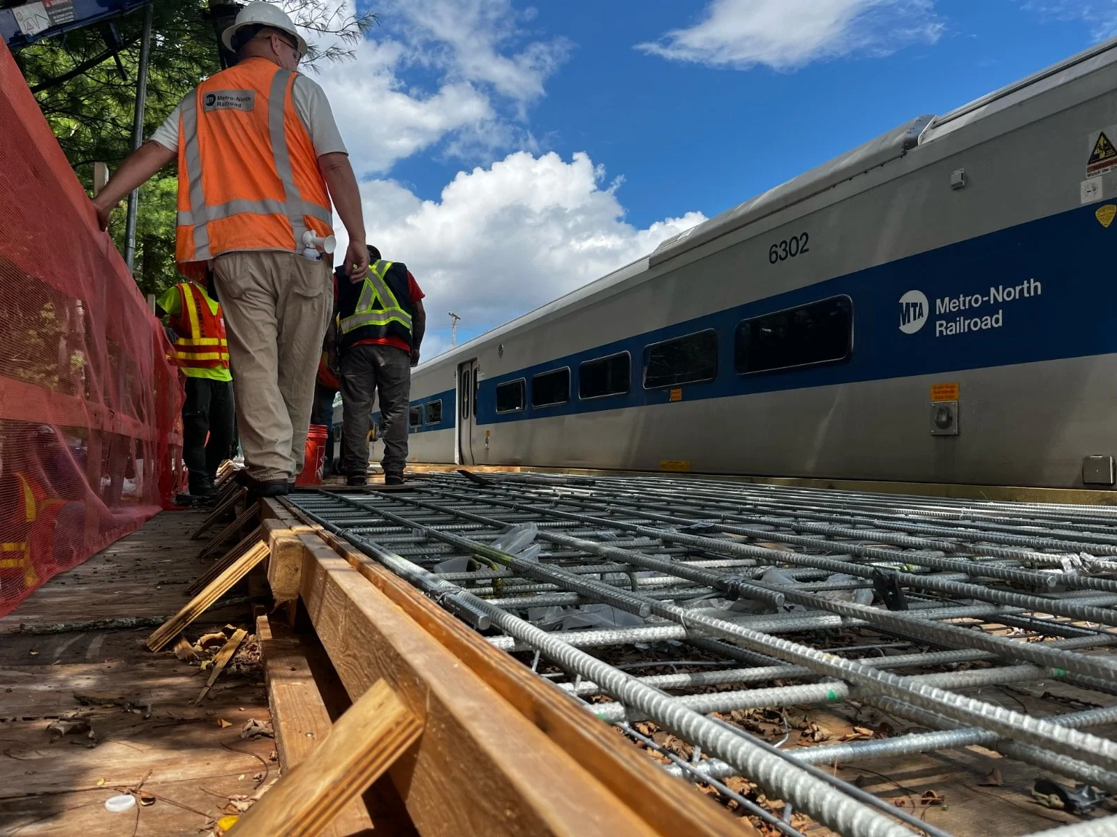 Construction workers standing near a parked Metro-North Railroad train on a track under a partly cloudy sky.
