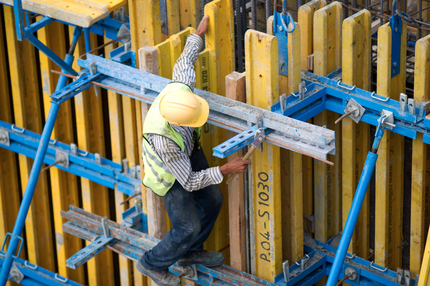 A construction worker wearing a yellow safety helmet and a yellow vest is installing or adjusting blue and yellow scaffolding on a construction site.