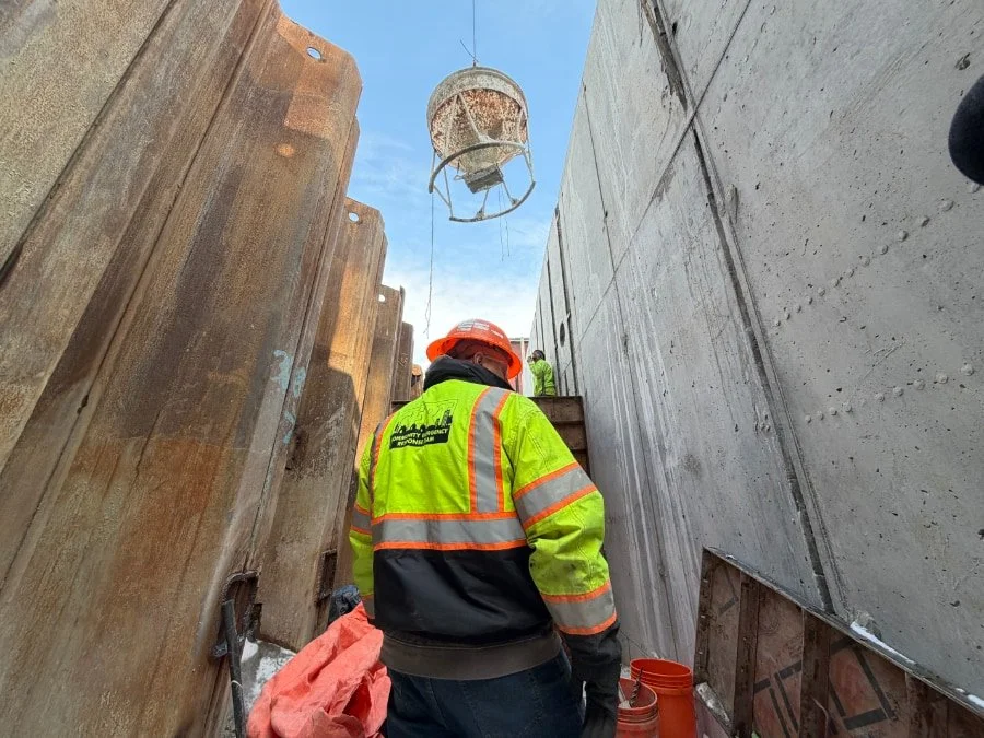 Construction worker wearing a high-visibility jacket and orange helmet at a construction site with concrete and wooden forms, a bucket suspended from above, and another worker in the distance.