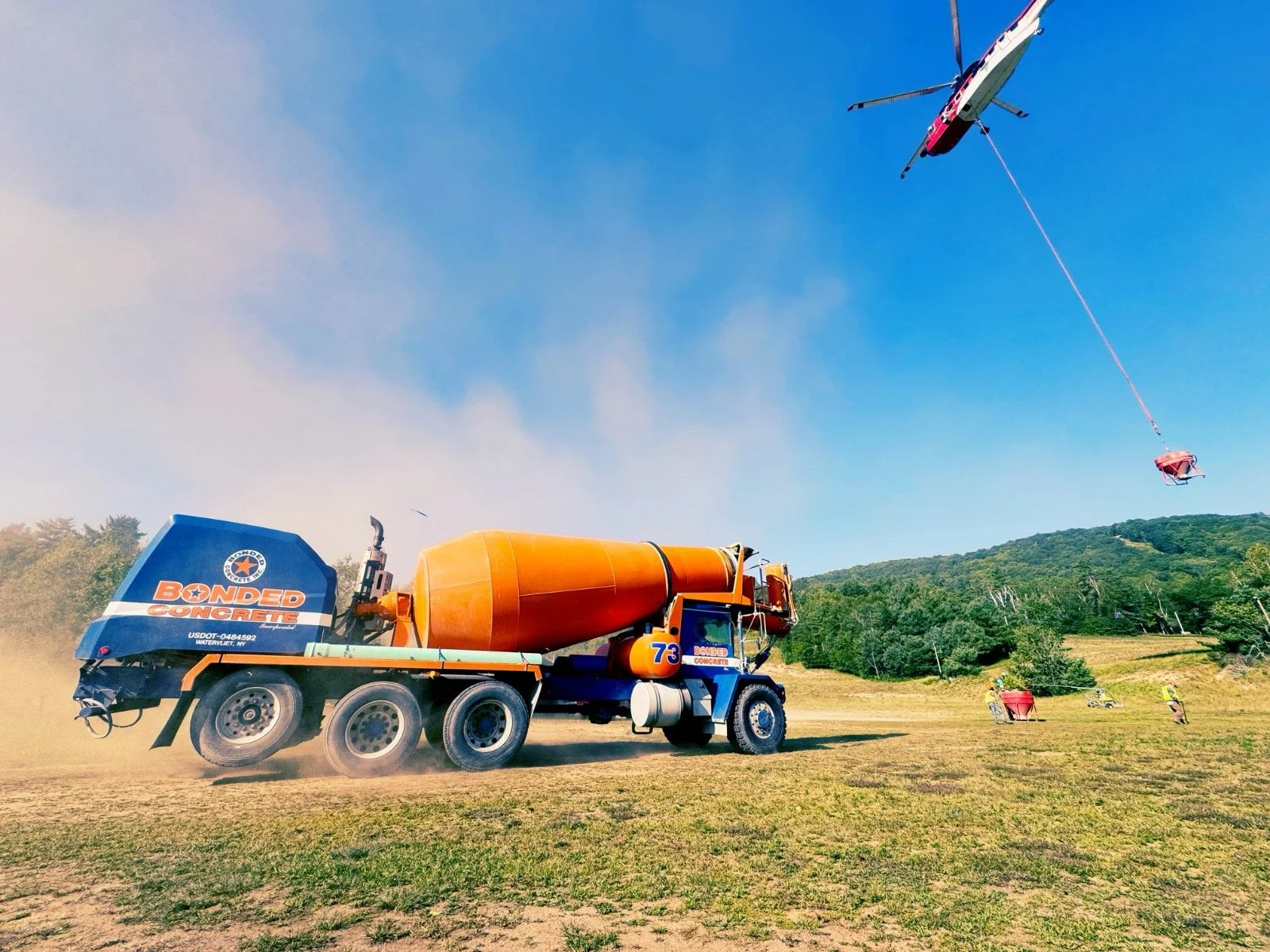 A construction site with a large orange and blue concrete mixer truck on a grassy field, and a helicopter flying overhead, hoisting a red crate via a cable. The background shows green hills and a clear blue sky.