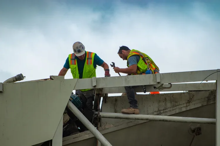 Two construction workers in safety vests and helmets working on a building's rooftop with cloudy sky background.