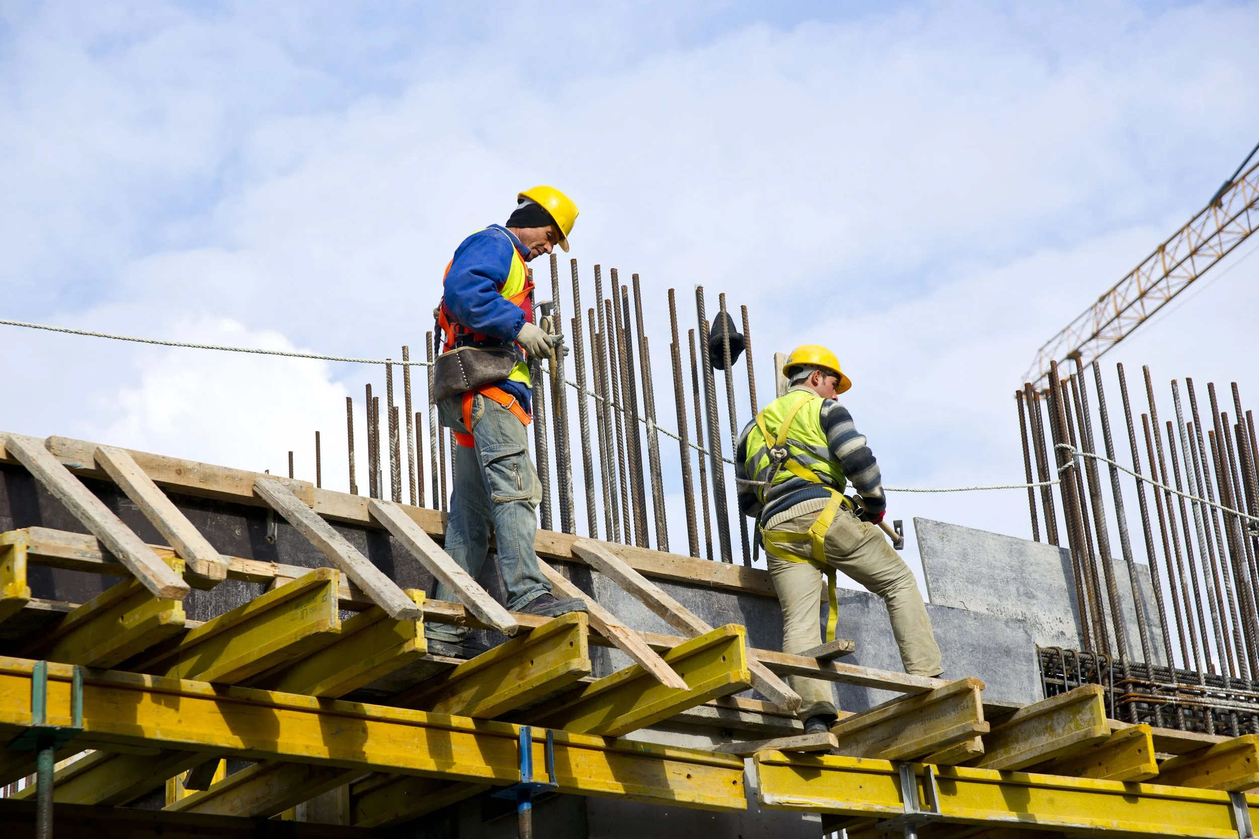 Two construction workers wearing yellow safety helmets and reflective vests working on a building under construction, with steel rebar and a blue sky in the background.