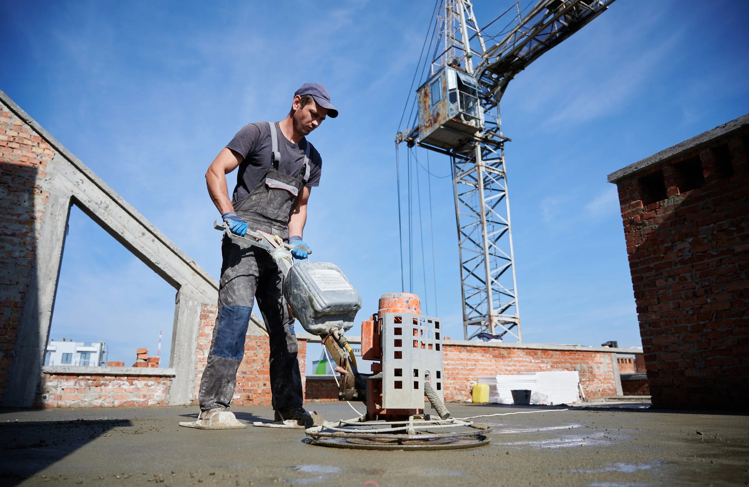 Construction worker operating a power tool on a concrete surface at a building site, with a crane and brick walls in the background.