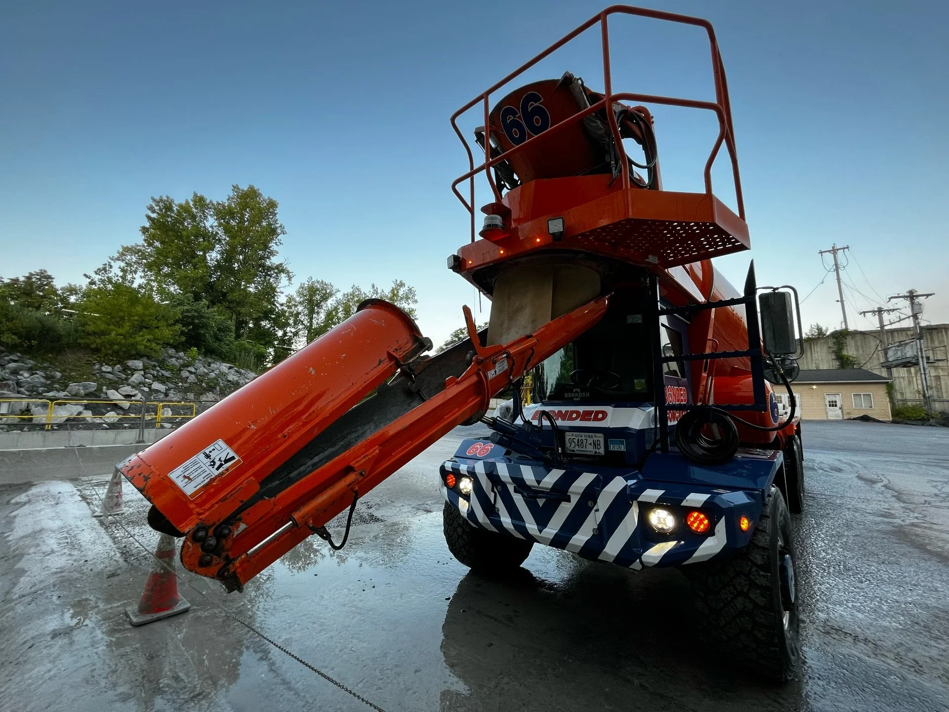 A bright orange street cleaning vehicle with an extended arm and chute, parked on a wet street under a clear sky, with trees and utility poles in the background.