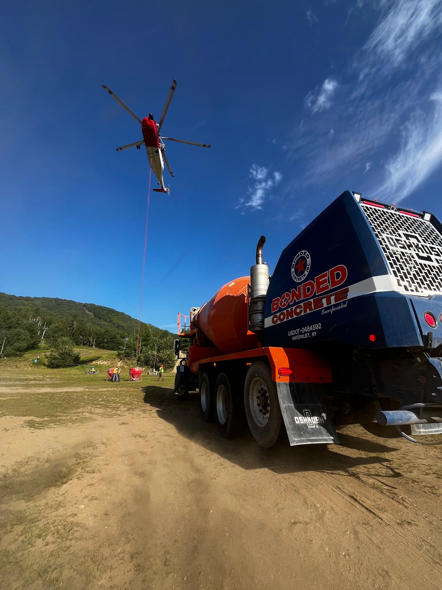 A construction site with a cement mixer truck and a helicopter hover with a cable, clear blue sky with some clouds, and distant green hills.