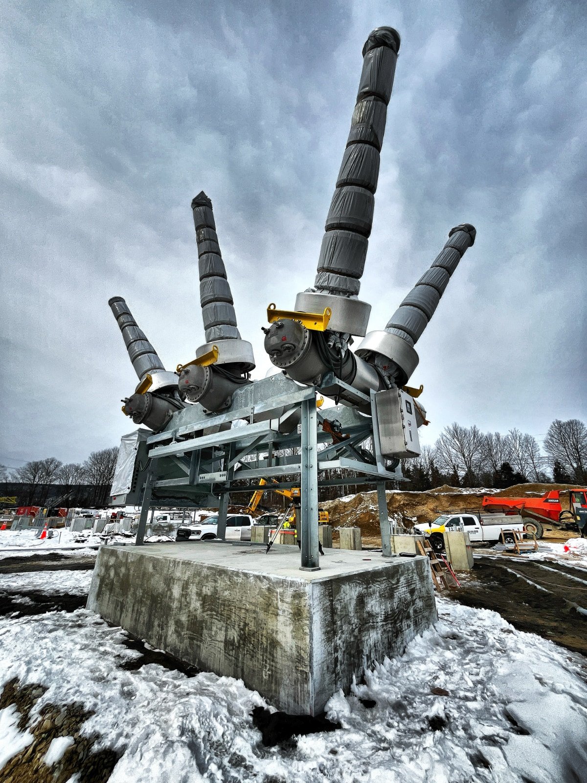 Large industrial transformer with four tall cooling fins on a concrete platform outdoors during winter, with snow on the ground and cloudy sky.