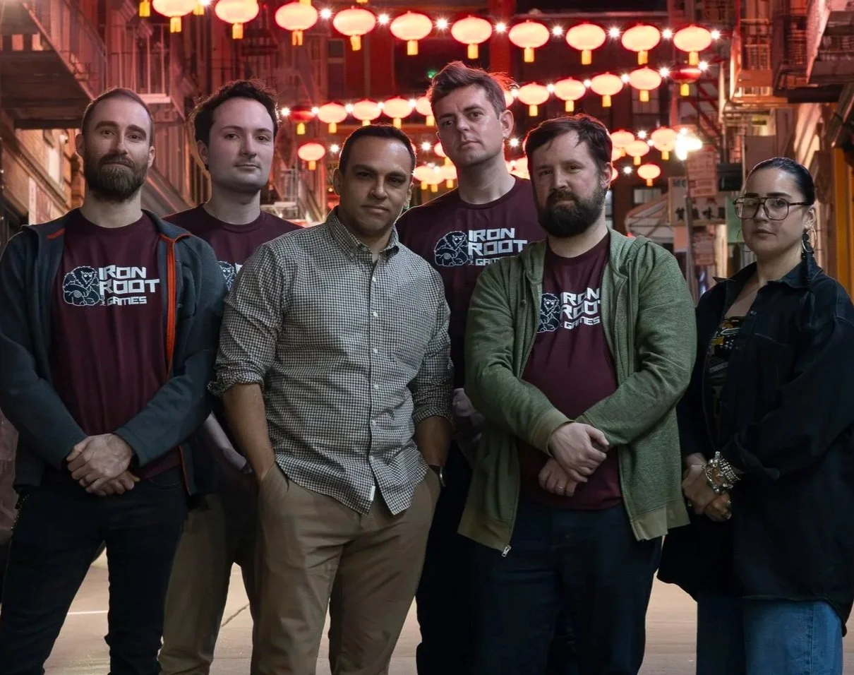 Group of six people standing together in front of a neon-lit street with hanging lanterns, some wearing matching 'Iron Root Games' shirts.