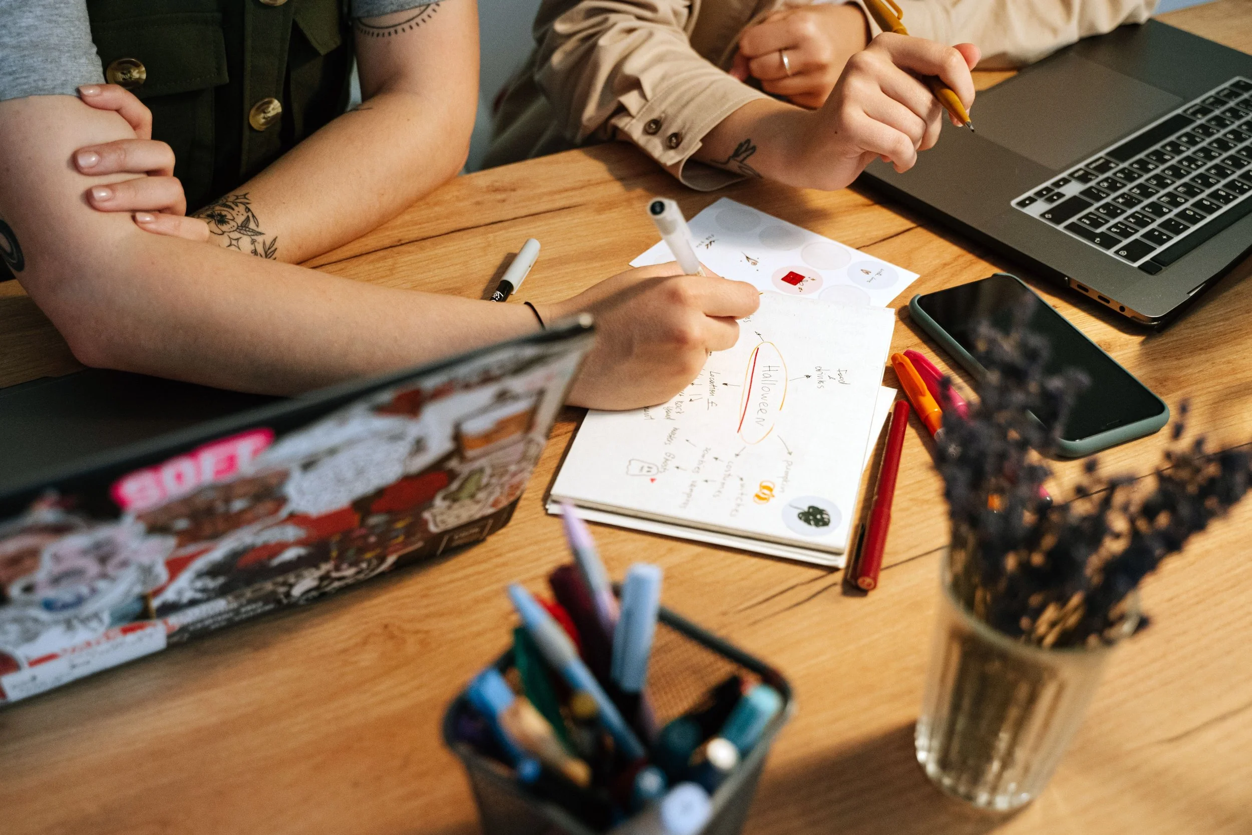 Two people sitting at a wooden table with various stationery items, a laptop, a smartphone, and notes. One person is writing, and the other has arms crossed.