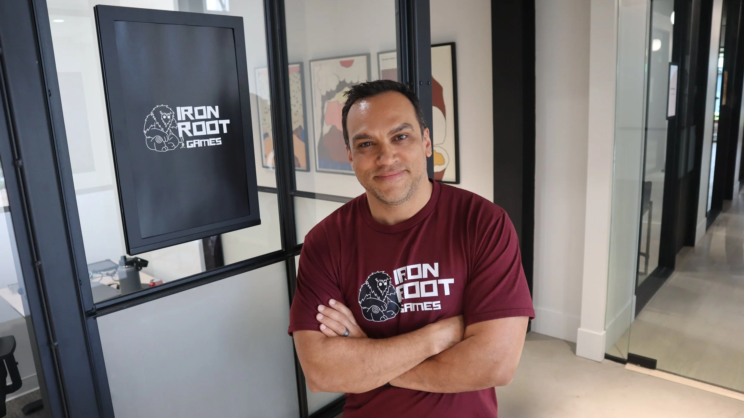 A man with arms crossed, wearing a maroon Iron Root Games T-shirt, standing in front of a computer screen with the same logo, in a modern office or gaming space.