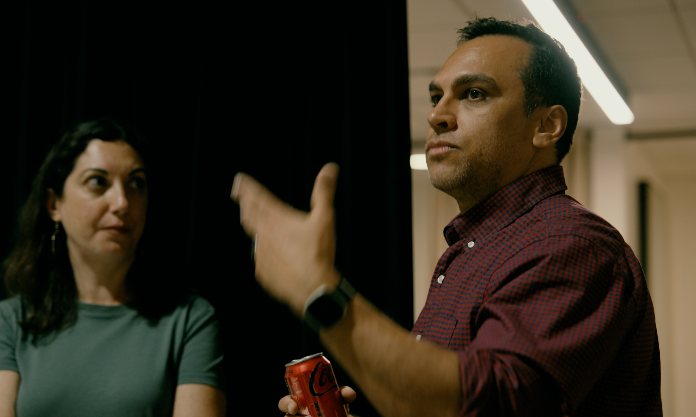 A man in a red checkered shirt holding a red soda can in front of an opaque black surface, with a woman in the background observing.