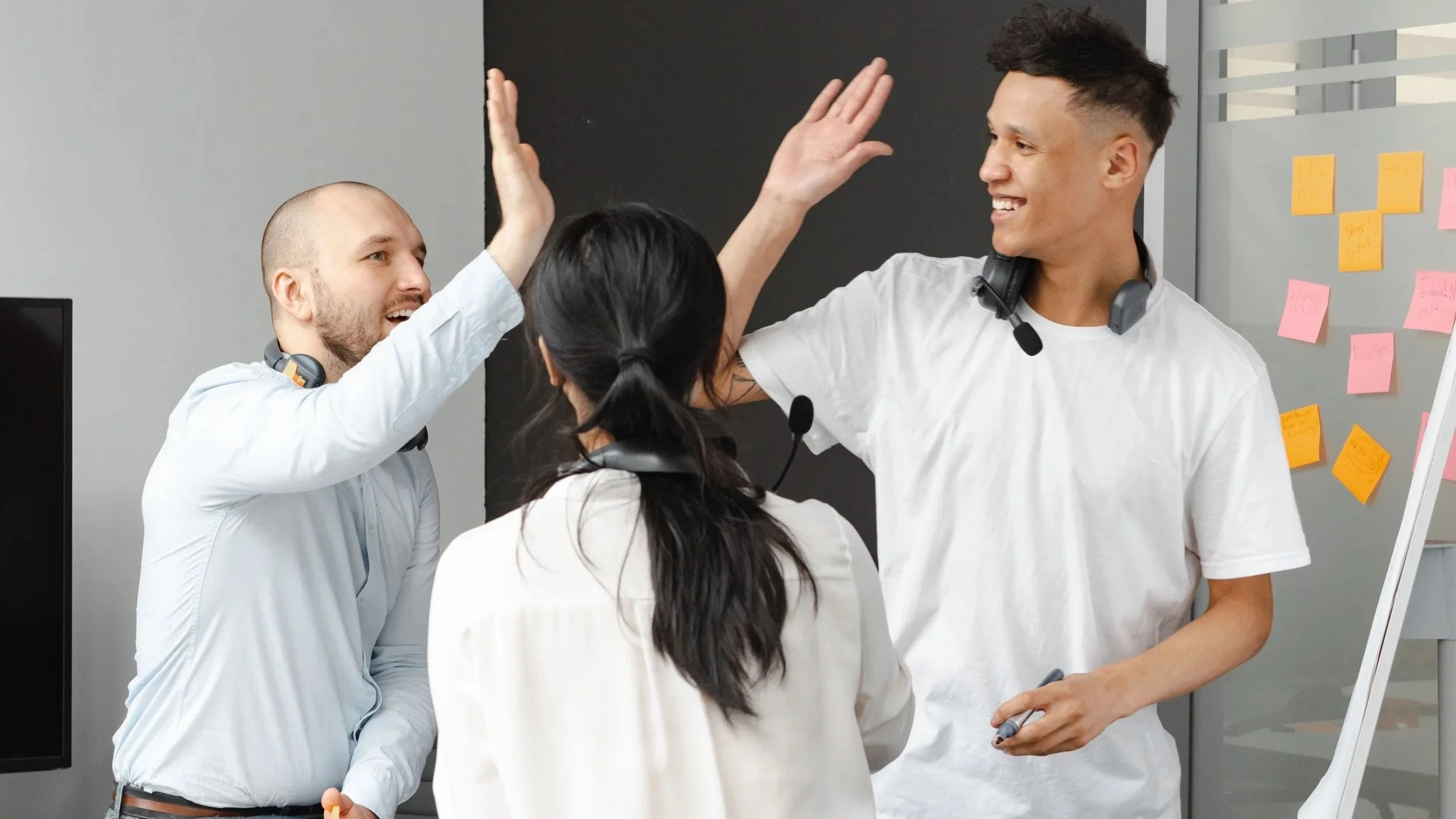 Three coworkers celebrating with high fives in an office, with colorful sticky notes on a whiteboard in the background.