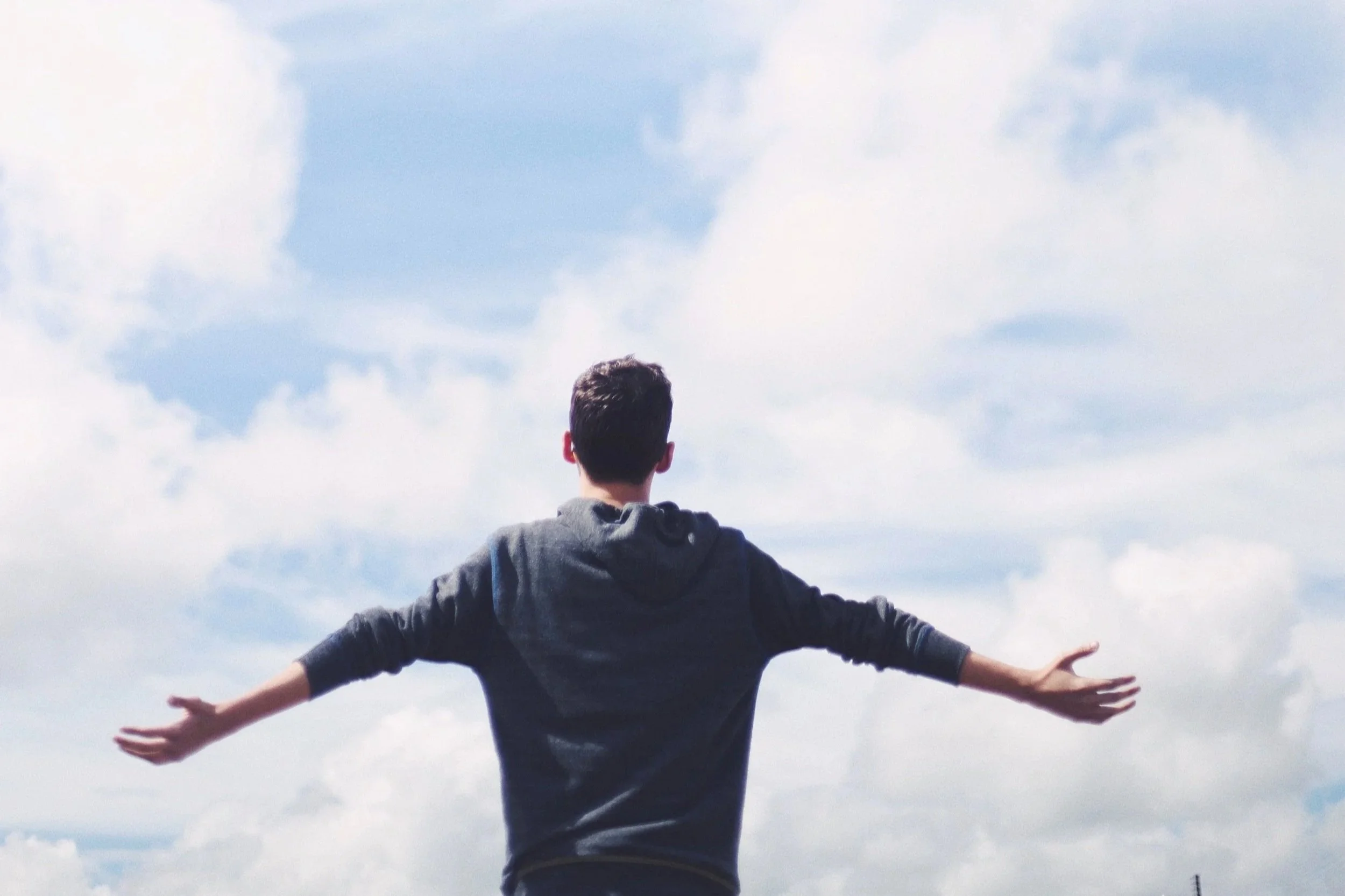 A person standing outdoors with arms extended outward and facing away from the camera, under a partly cloudy sky.