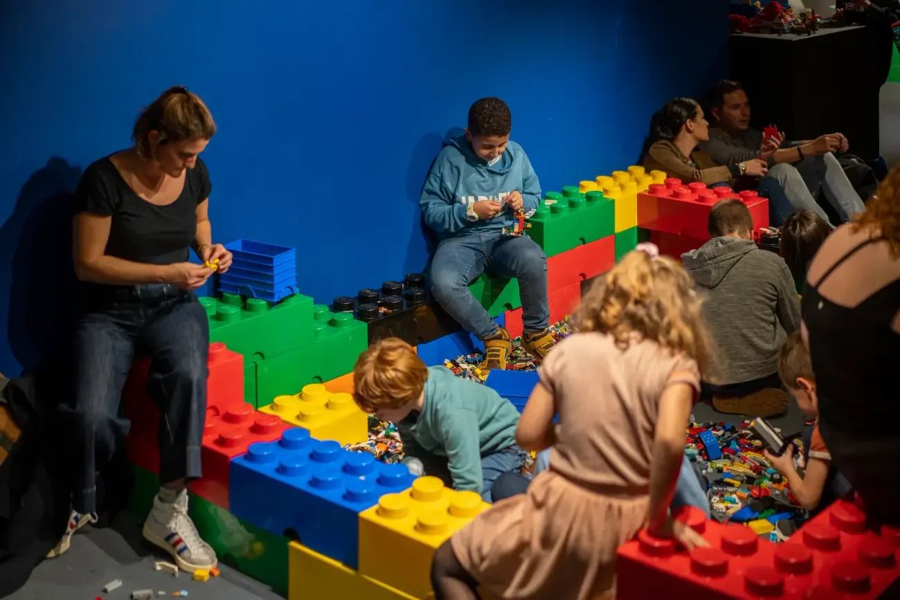 People sitting and playing with large LEGO bricks, building and arranging structures on the floor against a blue wall.