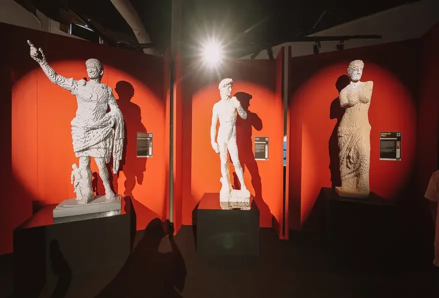 Three ancient Greek marble statues on display in a museum, each illuminated by spotlights against a red background, with informational plaques next to them.