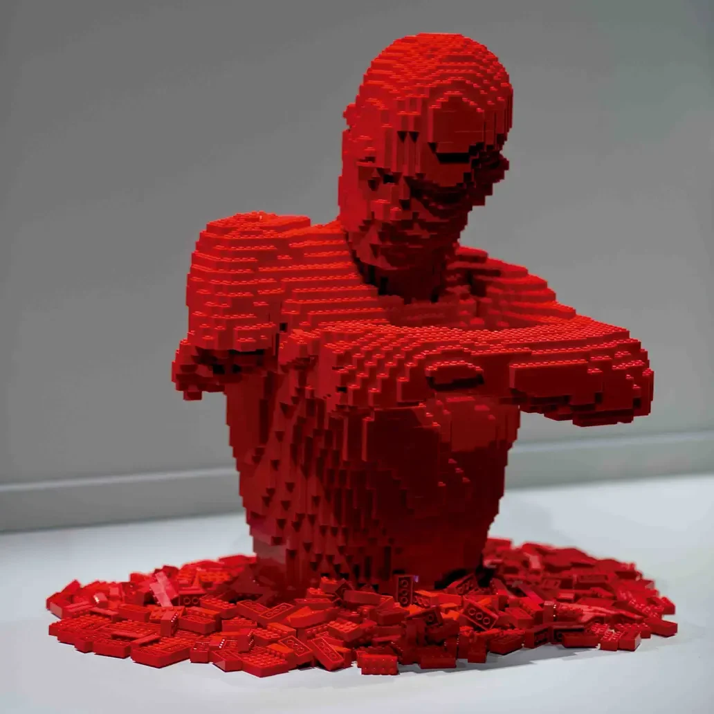 A sculpture of a woman made out of red LEGO bricks, sitting with her arms crossed and smiling, with LEGO pieces scattered around her base.