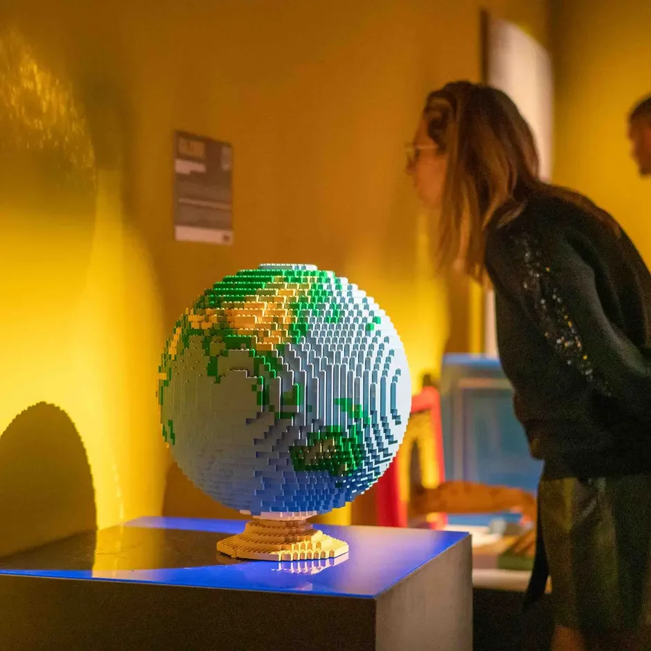 A life-sized model of the Earth made out of LEGO bricks, displayed on a black pedestal in a room with yellow walls, with a woman observing it.