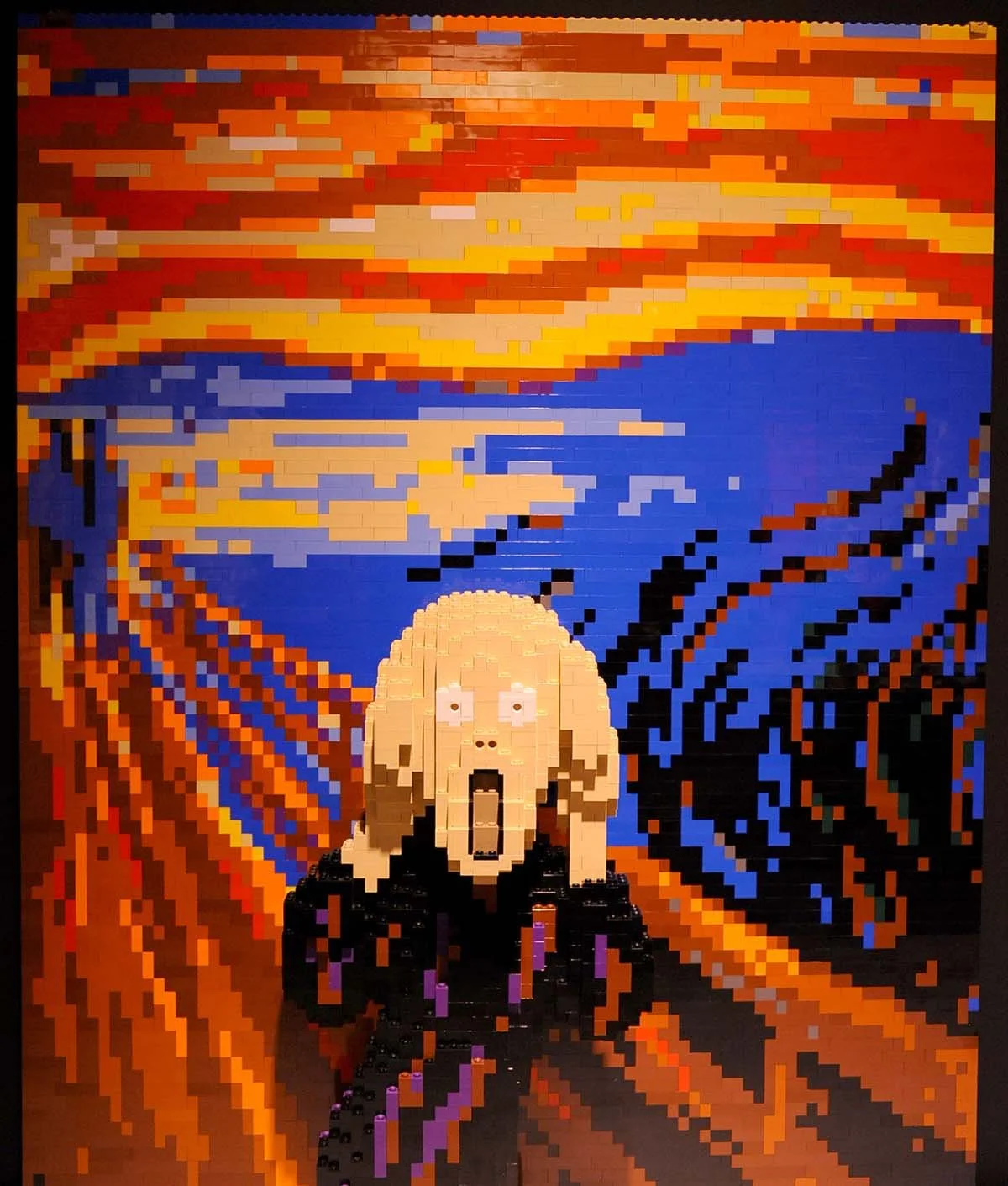 A large pixel art mural made of LEGO bricks depicting a woman with an open mouth in a scream, set against a vibrant background with swirling reds, oranges, blues, and yellows.
