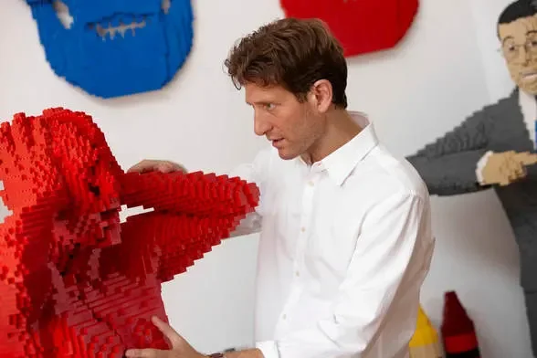 Man in white shirt examining a large red heart sculpture made of LEGO bricks in an indoor setting with art on the walls.