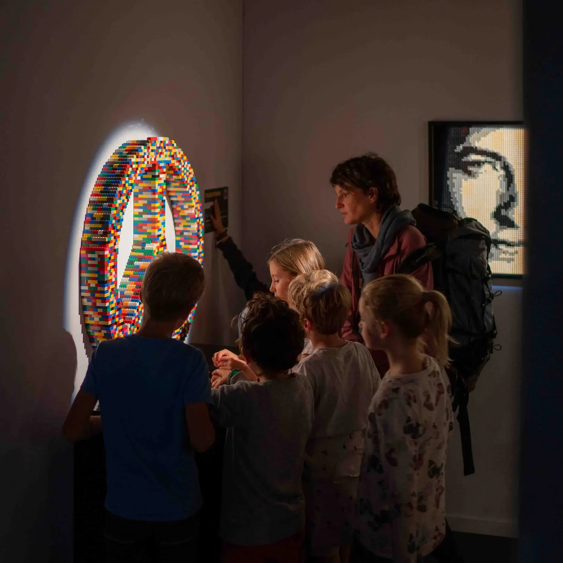 A group of children and a woman observing a large, wall-mounted LED artwork made of colorful LEGO bricks at a museum.