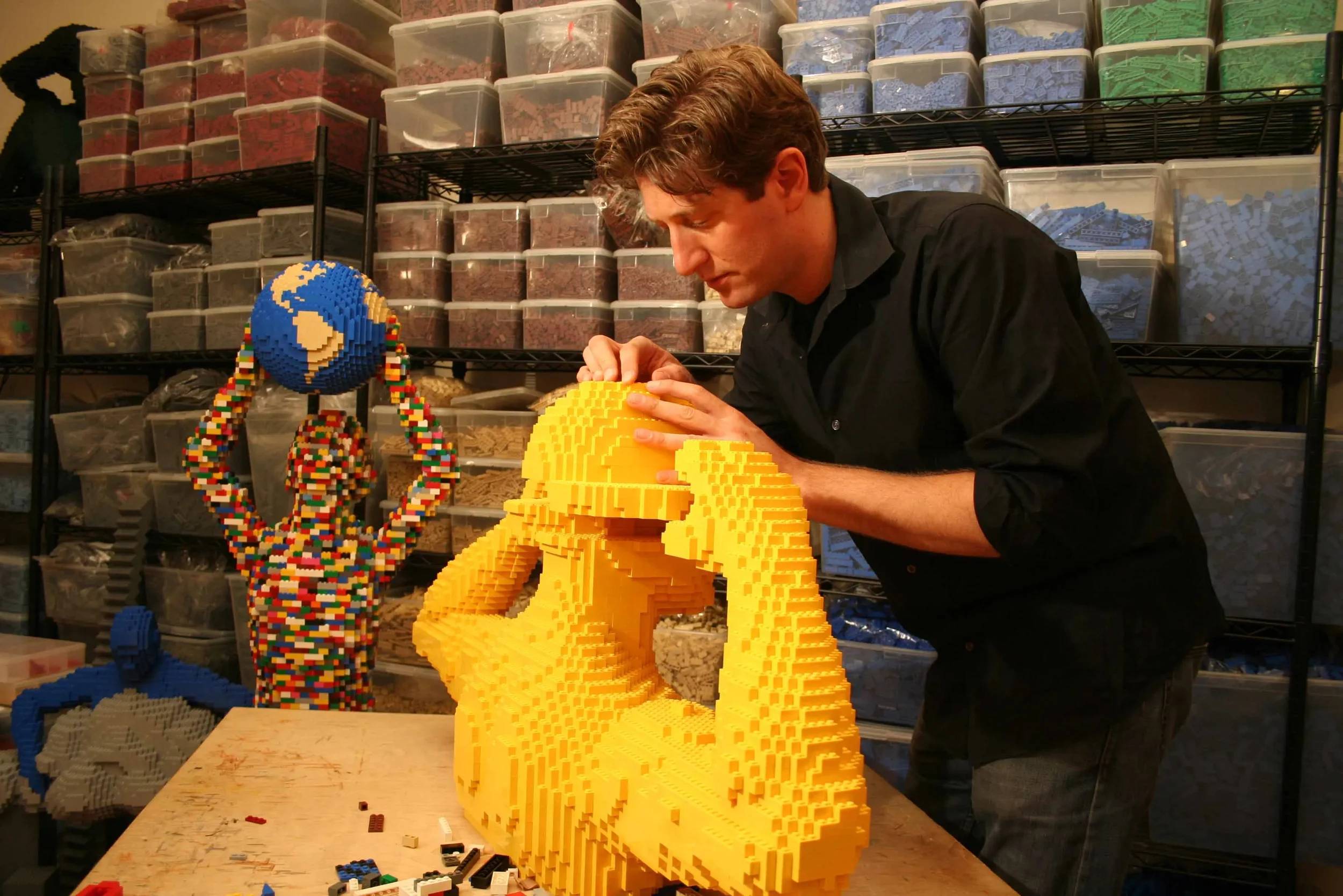 A man creating large LEGO sculptures, including a yellow piece and a colorful Earth figure, in a workshop surrounded by organized bins of LEGO pieces.