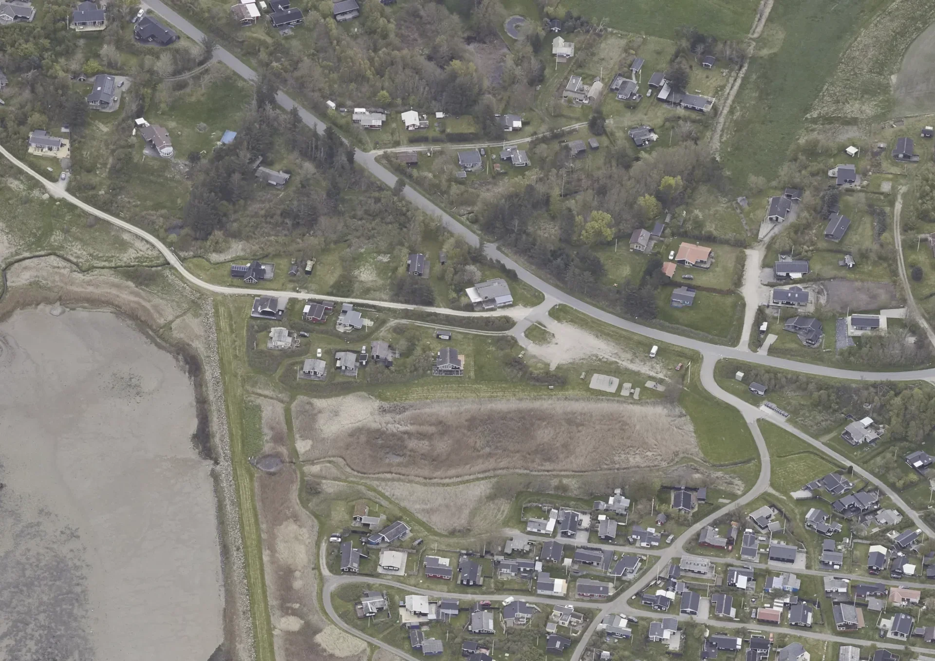 Luftfoto af et område med huse, veje og grønne områder, herunder en strand og et vand, taget fra luften.