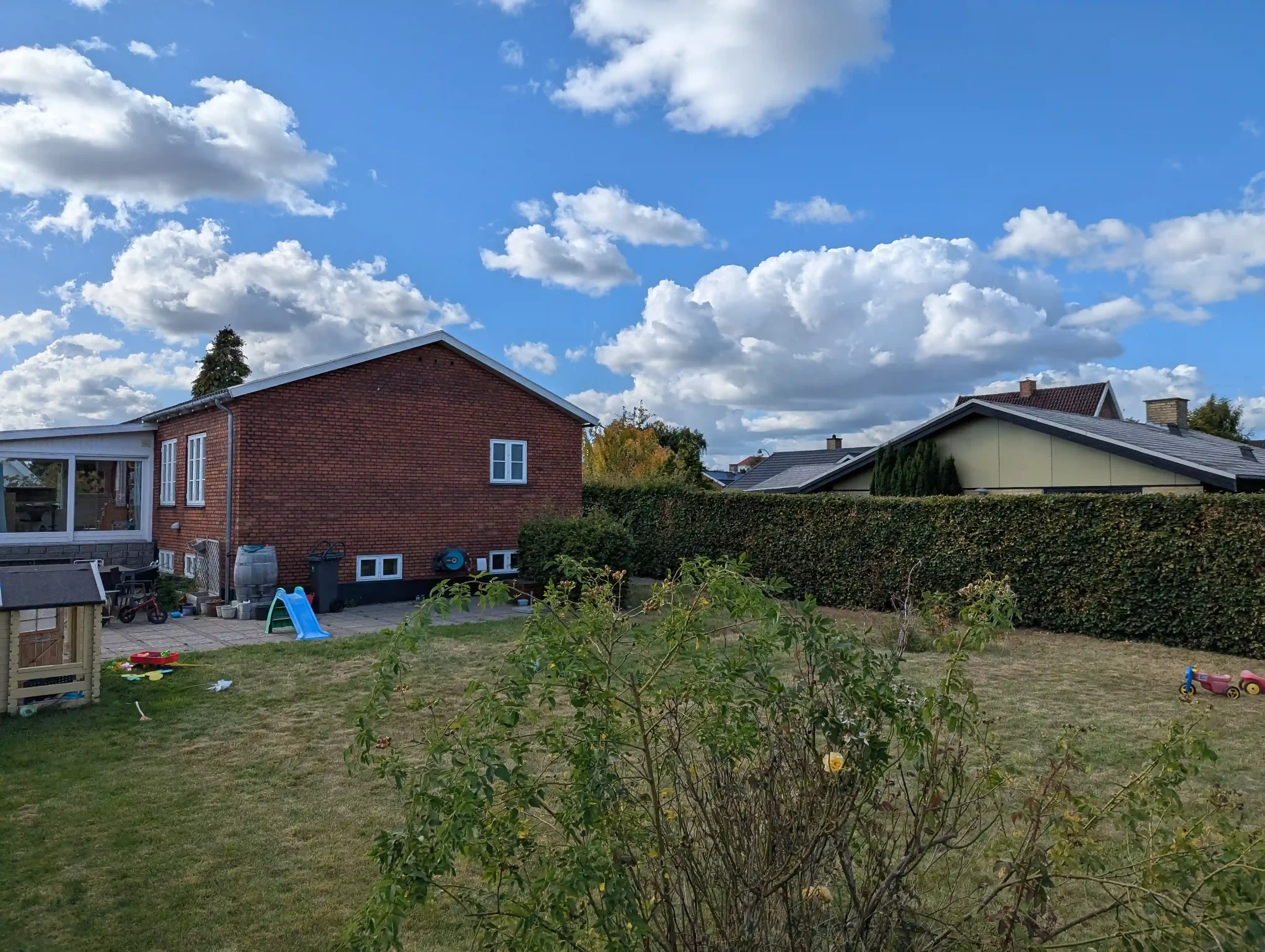 En baghave med græs, legetøj til børn, et rødbrunt hus med hvide vinduer, en hæk og en blå himmel med skyer.