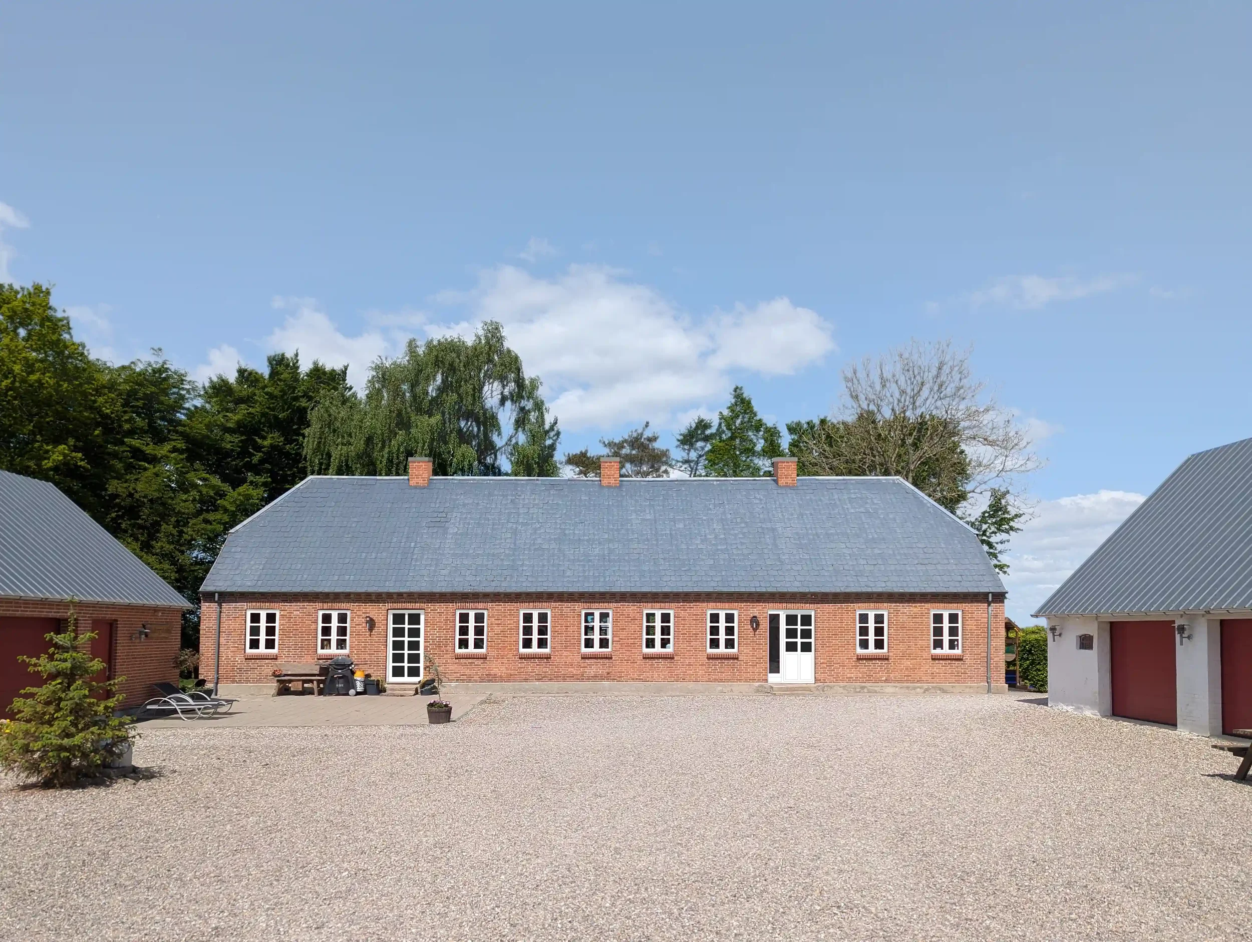 En rød-brick gård med blå tag og flere vinduer, omgivet af en stor grusplads, med to garagebygninger til højre og venstre, samt nogle sidde- og haveudstyr foran. Bagved er der grønne træer mod en blå himmel med enkelte hvide skyer.