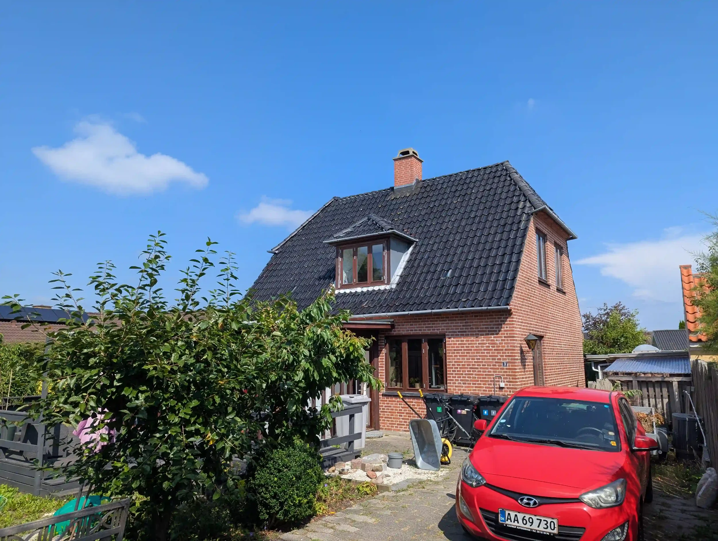 Et rødt murstenshus med sort tag, en stor vinduesbue på taget, og to mindre vinduer på siden. Der er en grøn busk foran huset, og en rød bil parkeret ude foran.