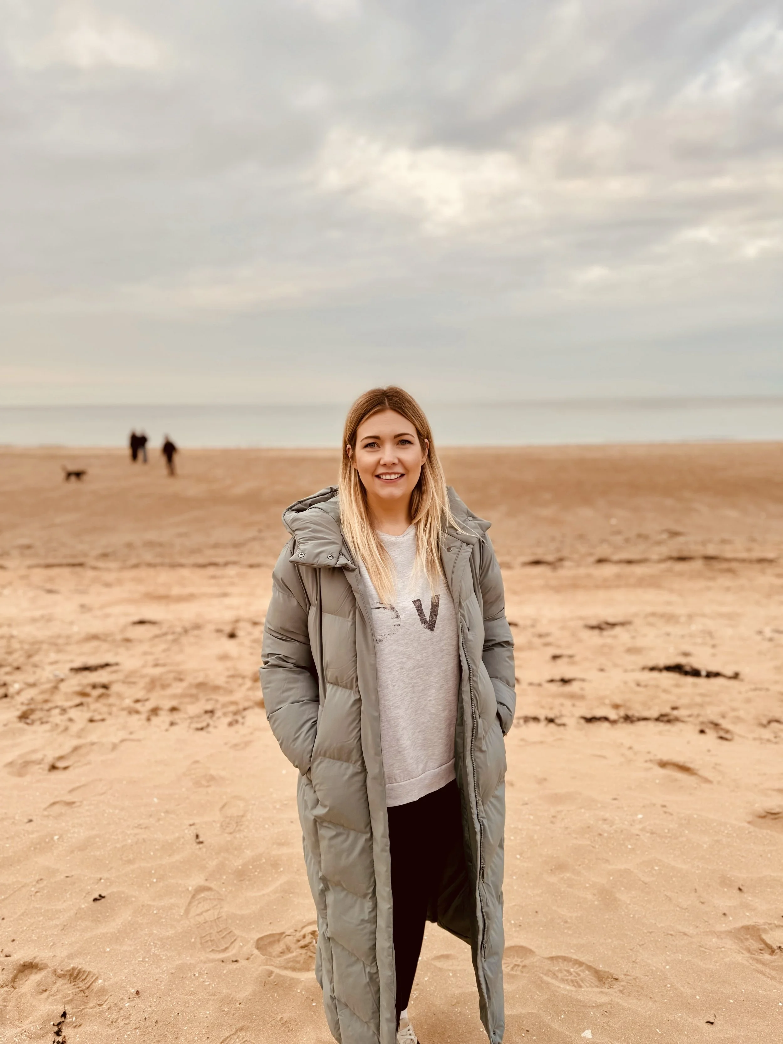 Young woman standing on a sandy beach with a cloudy sky, wearing a puffy gray coat and smiling at the camera.